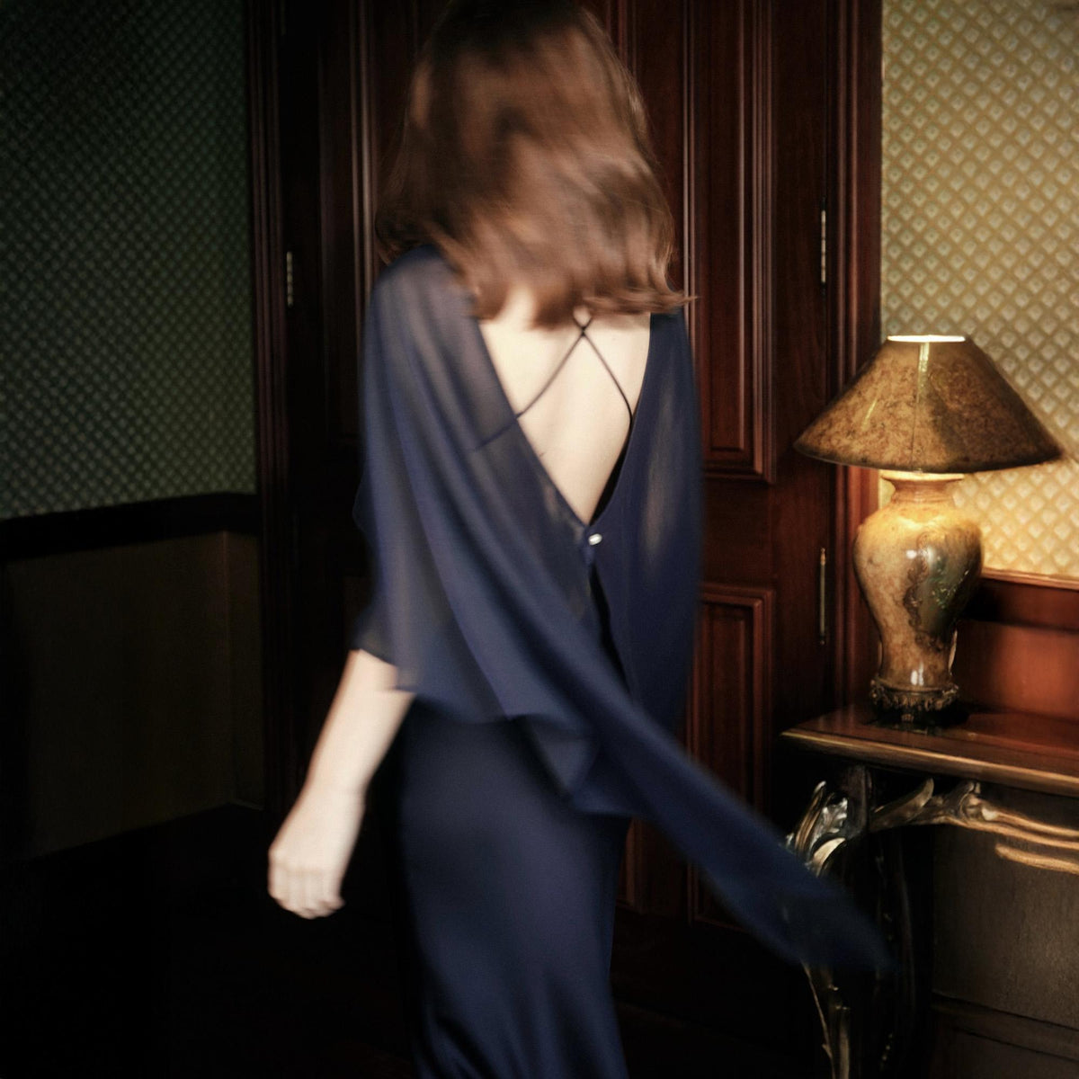 May include: A dark navy blue dress featuring a deep open back design with thin crisscross straps and a flowing, sheer cape-like overlay that extends from the shoulders. The garment has a smooth, fitted silhouette and a single visible button closure.