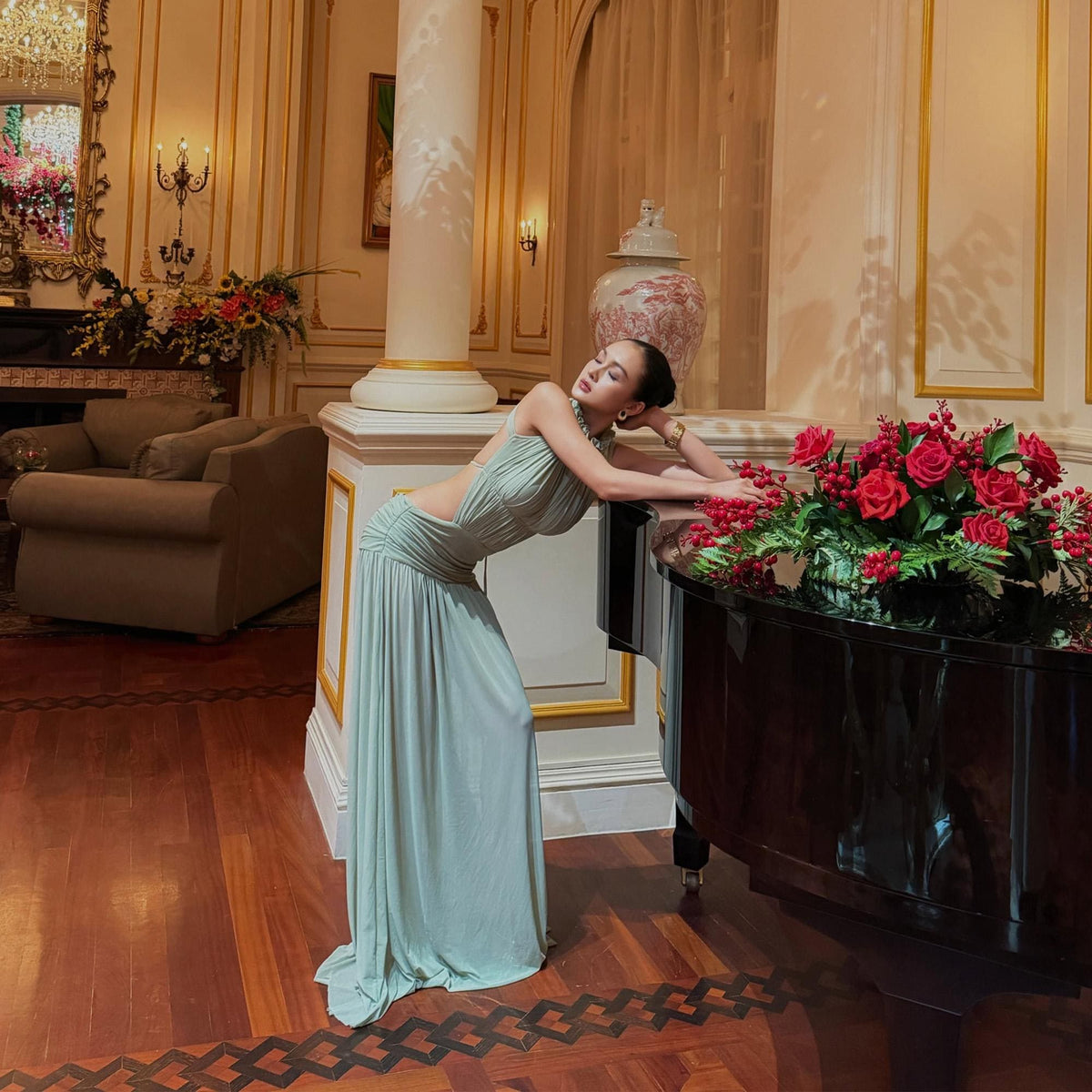 May include: A floor-length, light sage green sleeveless gown featuring a ruched bodice, a high neckline, and an open back design. The dress is crafted from a soft, flowing fabric that drapes to the floor, creating a fluid silhouette with a gathered waistline.