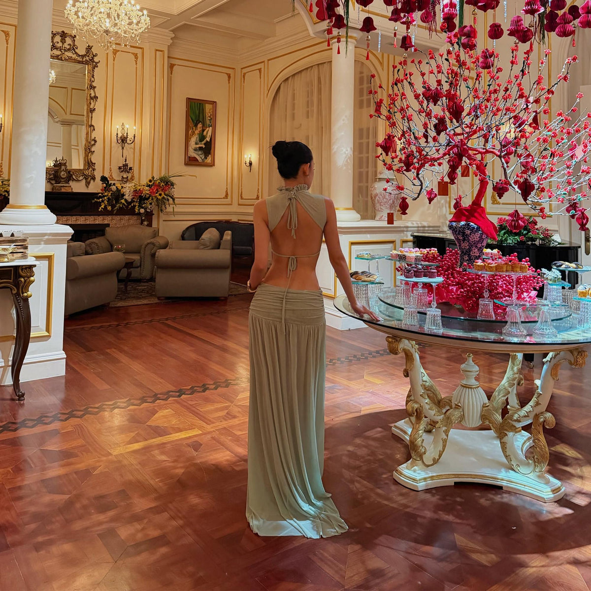 May include: A sage green floor-length dress featuring a sleeveless halter-style bodice with a high neckline and an open back design. The skirt has a gathered, ruched waistline that flows into a long, straight silhouette, creating a draped effect.