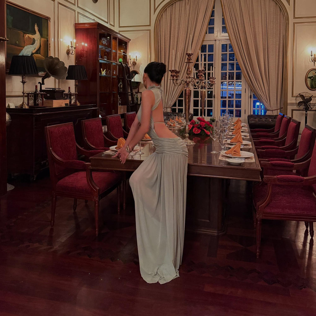 May include: A floor-length, light sage green sleeveless dress featuring a high neckline, an open back design, and gathered fabric detailing at the waist that flows into a long, straight skirt. The garment is made of a smooth, lightweight material.