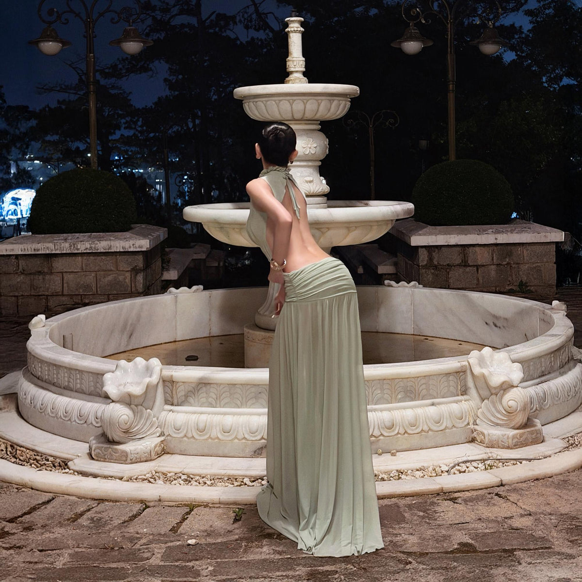 May include: A floor-length sage green dress featuring a high halter neckline, an open back design, and a ruched waistline that transitions into a flowing, pleated skirt. The garment is crafted from a lightweight fabric that drapes to the floor.