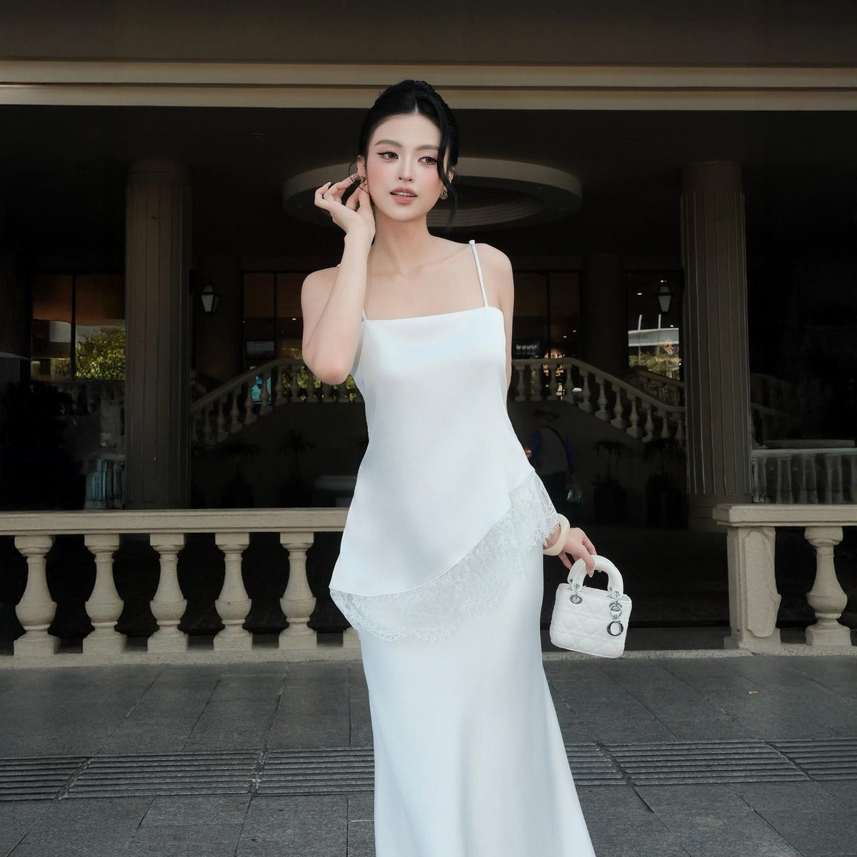 May include: A floor-length white slip dress with thin spaghetti straps and a square neckline. The dress features an asymmetrical hemline accented with a delicate white lace trim. A small, white, quilted handbag with silver hardware is held.