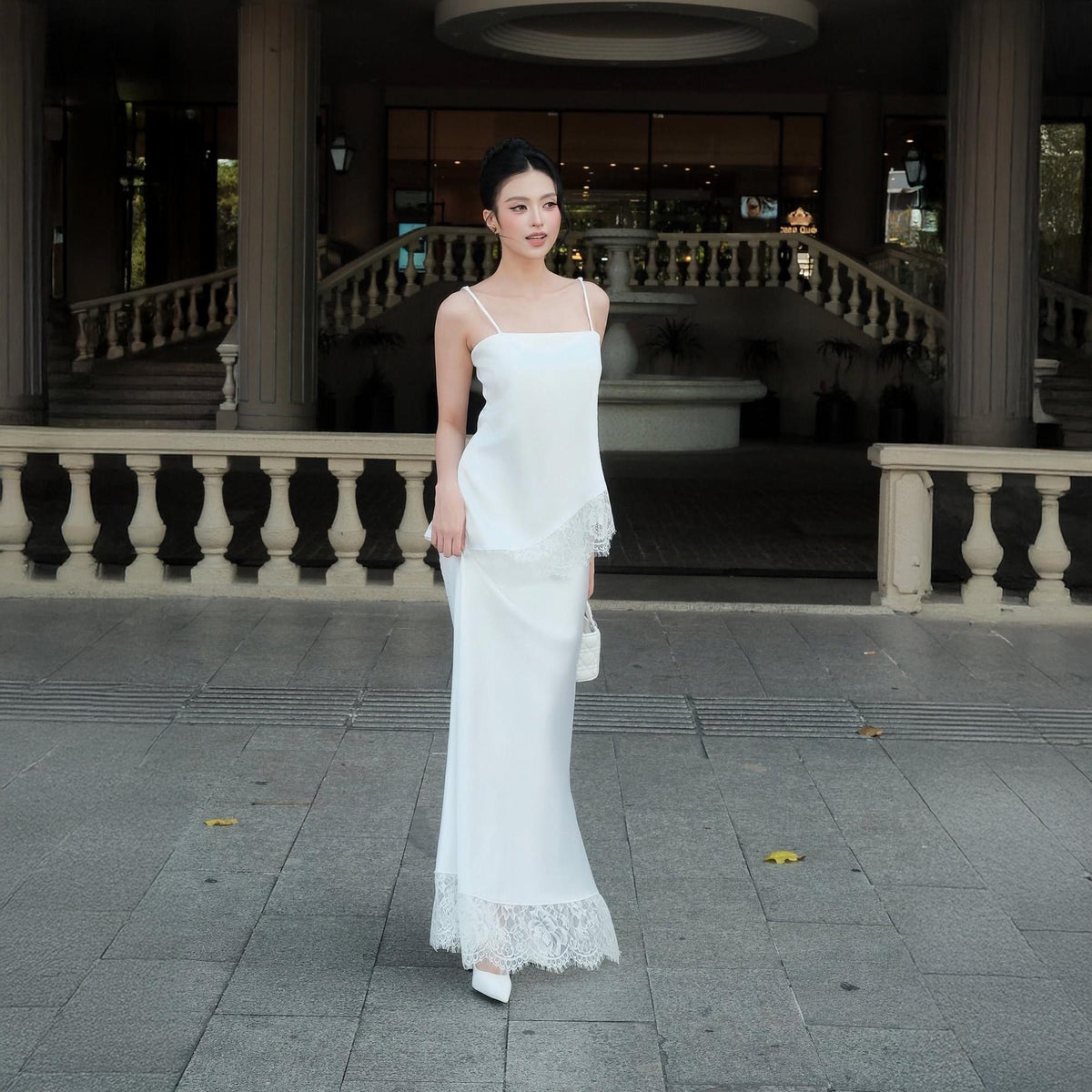 May include: A floor-length white dress featuring thin spaghetti straps, a straight neckline, and a smooth bodice. The garment includes an asymmetrical overlay with delicate lace trim at the hem and a matching lace border along the bottom edge.