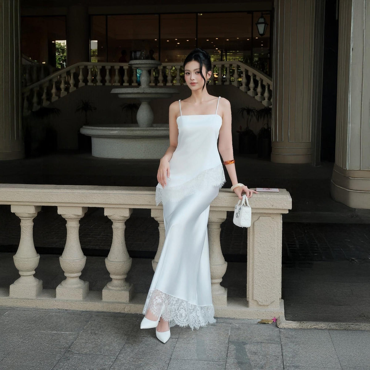 May include: A floor-length white satin slip dress featuring thin spaghetti straps, a straight neckline, and an asymmetrical lace overlay detail at the hip. The hemline is finished with a wide band of intricate white lace and matching white heels.