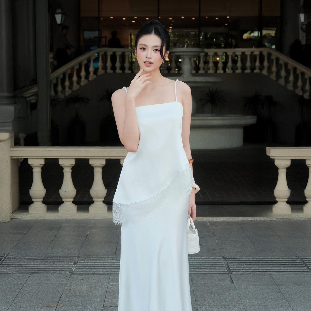 May include: A floor-length white dress featuring thin spaghetti straps, a straight neckline, and an asymmetrical hemline accented with delicate white lace trim. The garment has a smooth, flowing silhouette and is paired with a small white handbag.