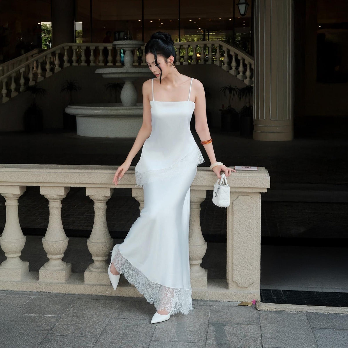 May include: A floor-length white satin slip dress featuring thin spaghetti straps, a straight neckline, and a layered bodice design. The hemline is finished with a wide, intricate white lace trim. The dress is paired with white pointed-toe heels.