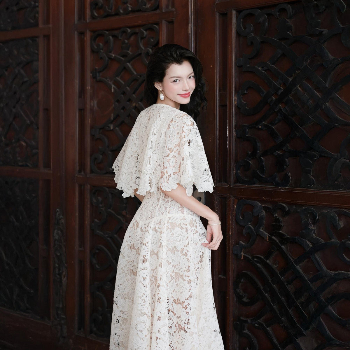 May include: A white lace dress featuring a short-sleeved capelet overlay with a scalloped hemline. The garment is constructed from intricate floral lace fabric that extends from the shoulders down to a flowing skirt with a defined waistline.