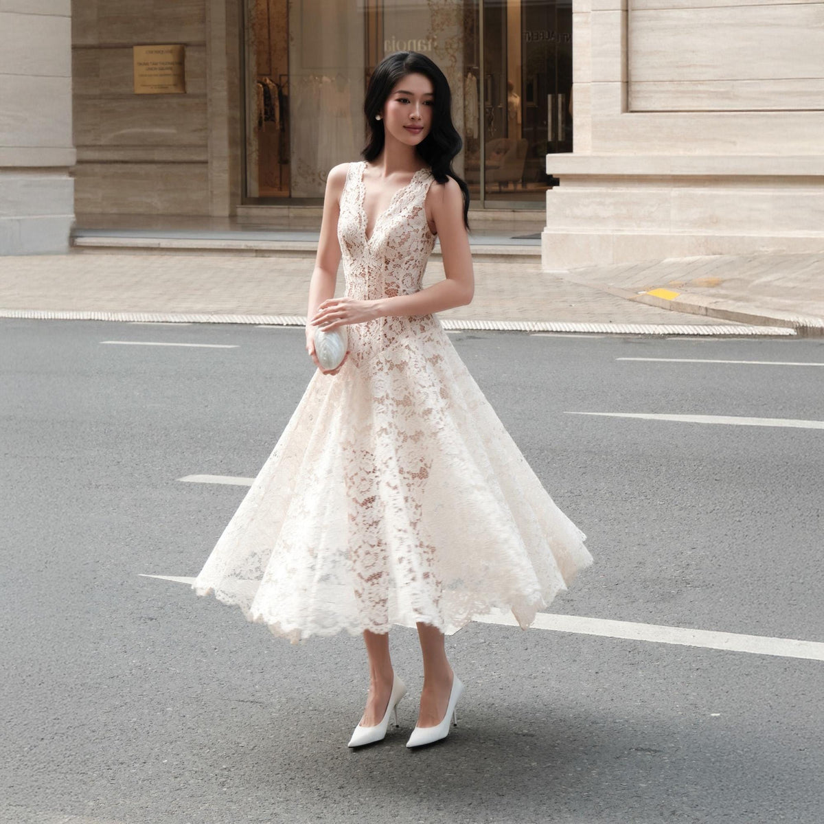 May include: A sleeveless midi-length dress made of cream-colored lace with a deep V-neckline and a flared skirt. The dress features a fitted bodice and a scalloped hemline. It is paired with white pointed-toe high-heeled pumps and a small clutch.