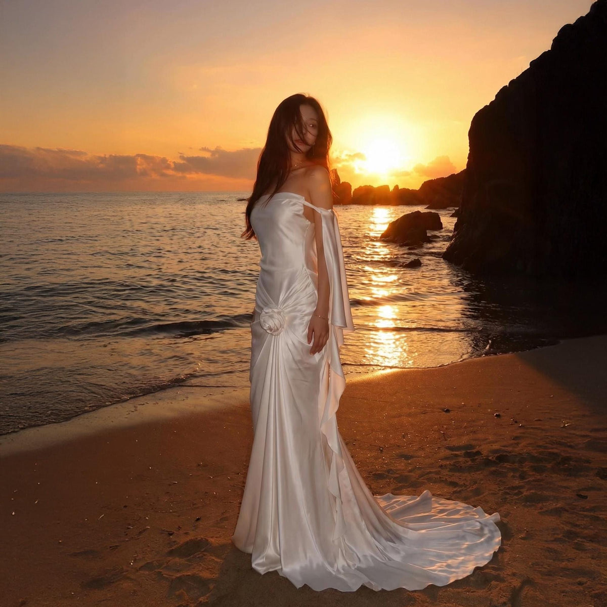 May include: A floor-length white satin gown featuring a strapless bodice, a gathered waist detail with a central fabric flower, and long, flowing detached sleeves that drape from the shoulders, ending in a slight train on the sandy beach.