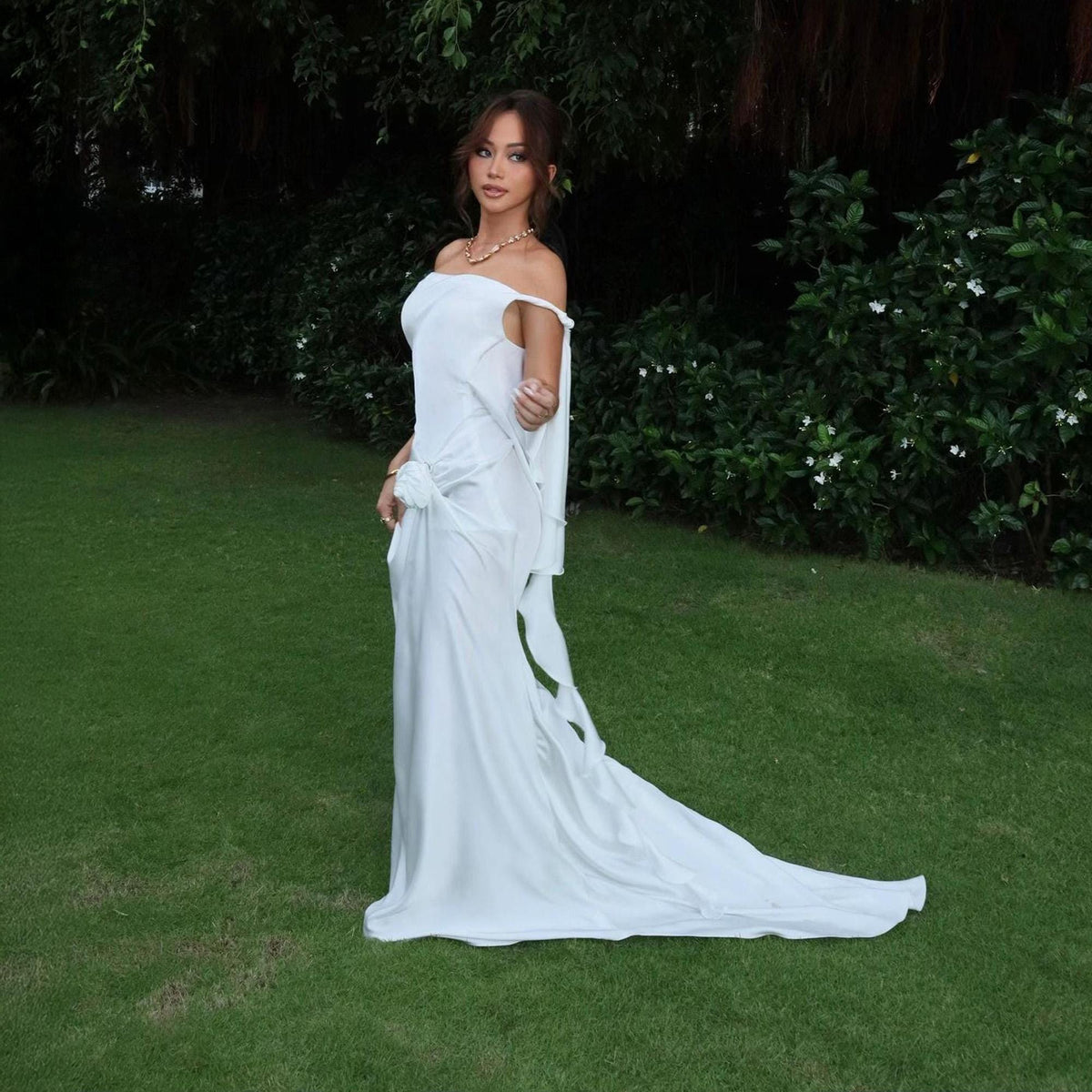 May include: A floor-length white off-the-shoulder gown featuring a draped bodice, a gathered fabric detail at the waist, and a flowing skirt with a train. The garment is constructed from a smooth, satin-like fabric with a subtle sheen.