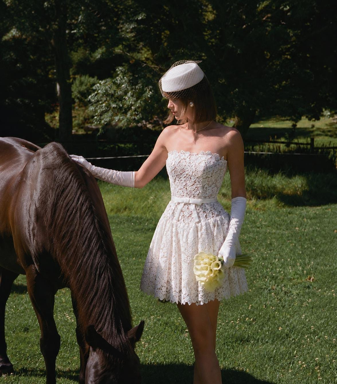May include: A strapless white lace mini dress with a fitted bodice, a thin waist belt, and a flared skirt. The ensemble includes long white gloves, a white pillbox hat with a brown netting veil, and a bouquet of white calla lilies held in one hand.