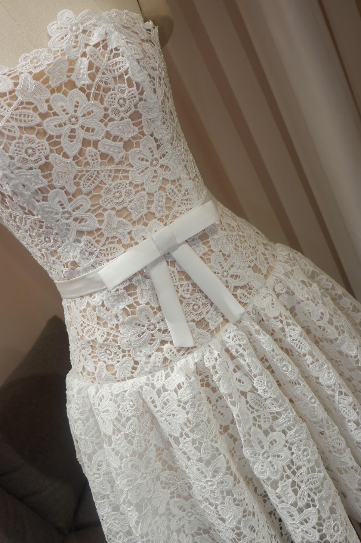 May include: A white strapless dress featuring an intricate floral lace overlay pattern throughout the bodice and skirt. A smooth, solid white satin ribbon is tied into a bow at the waistline, contrasting with the textured lace fabric.