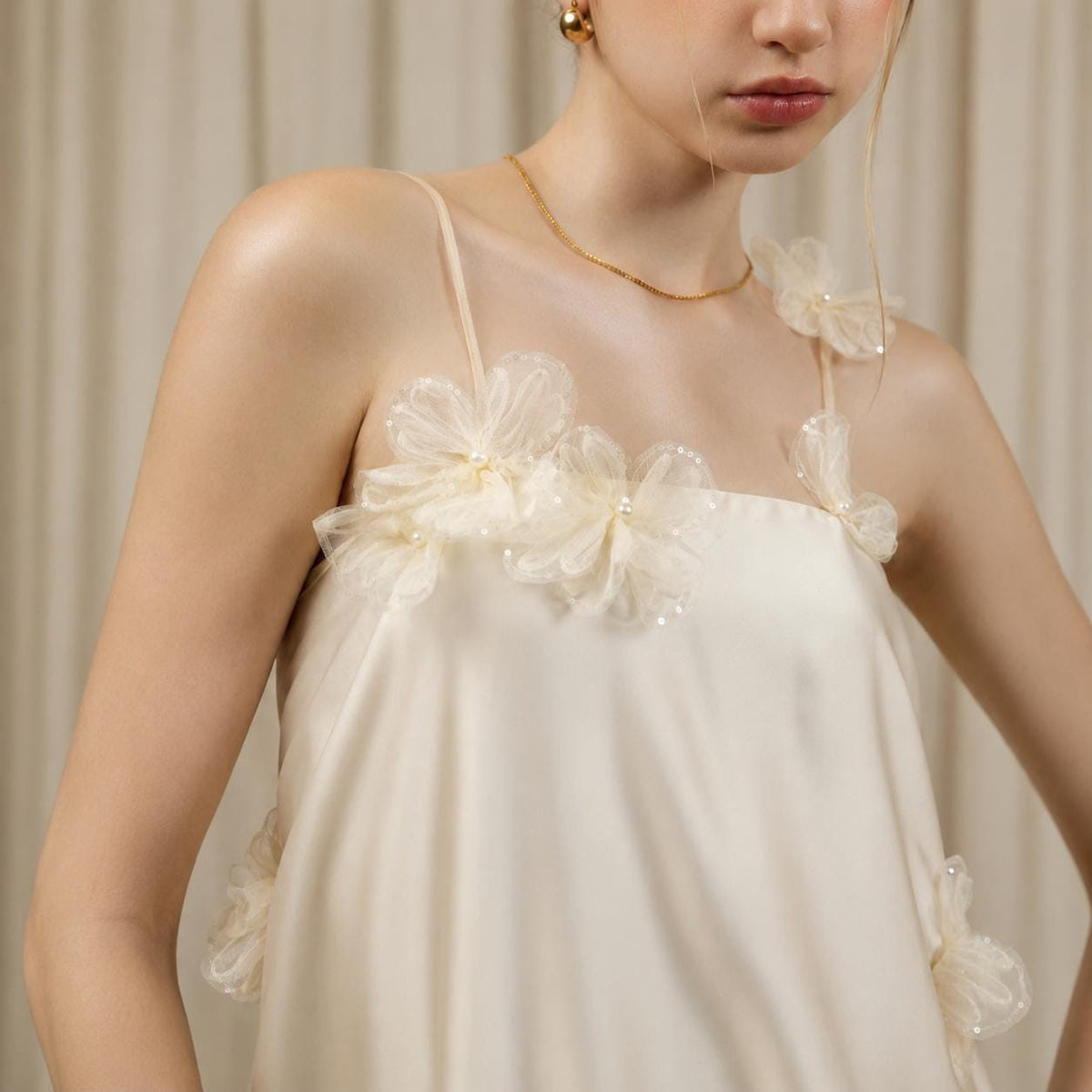 May include: A cream-colored sleeveless top featuring thin spaghetti straps and layered sheer fabric flowers attached to the neckline and straps. Each flower is accented with a small white pearl at the center, creating a textured floral detail.