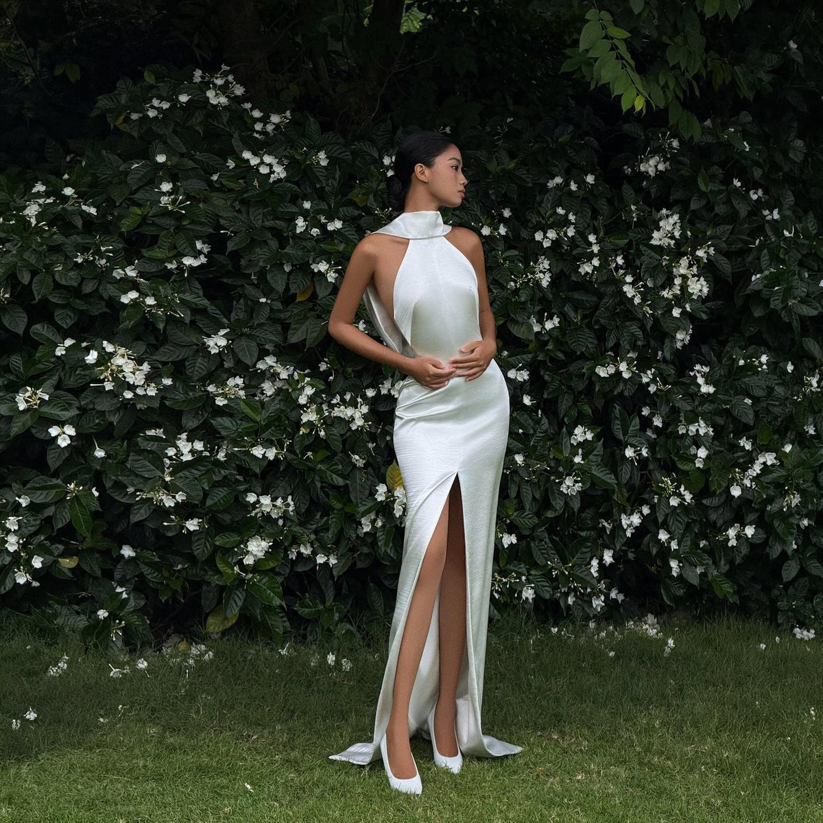 May include: A floor-length white satin gown featuring a high halter neckline, an open back design, and a prominent front slit that extends from the mid-thigh to the hem. The dress is paired with matching white pointed-toe heels.