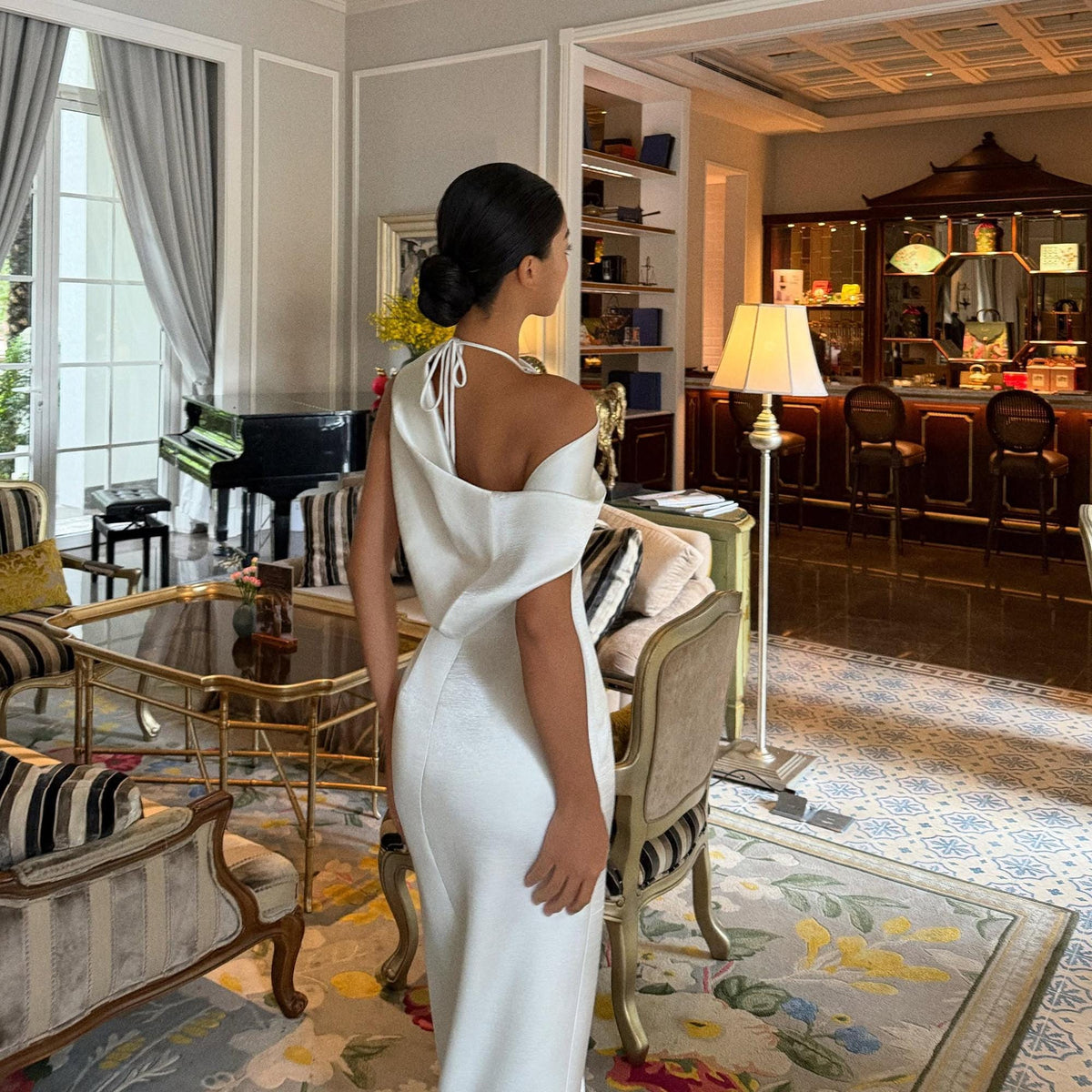 May include: A floor-length white dress featuring a draped, asymmetrical neckline with thin spaghetti straps that tie at the back. The garment has a fitted silhouette and a smooth, solid-colored fabric construction, showcasing a clean and simple design.