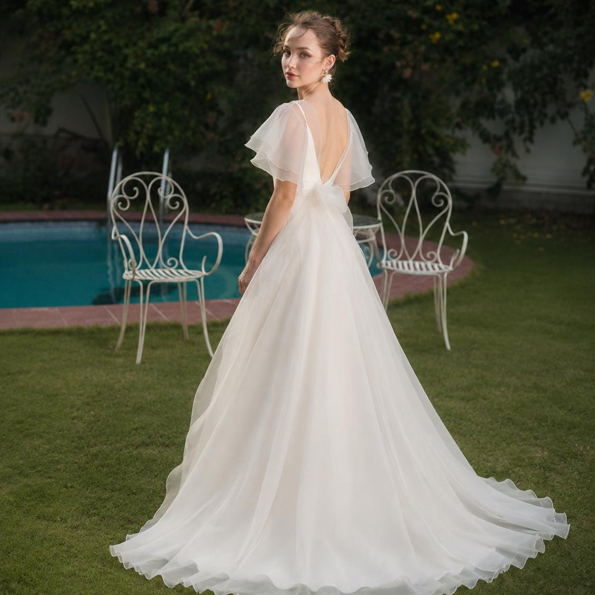 May include: A floor-length white wedding dress featuring a deep V-back, sheer flutter sleeves, and a flowing A-line skirt. The gown is constructed with a fitted bodice that transitions into a full, layered skirt that pools slightly on the ground.