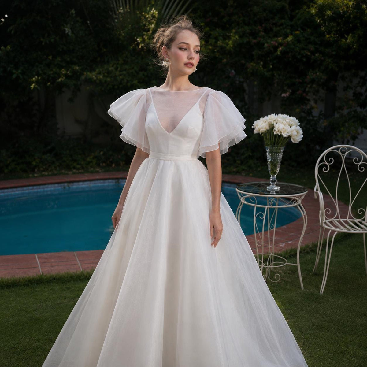 May include: A floor-length white gown featuring a deep V-neckline, a sheer illusion bodice, and layered tulle flutter sleeves. The dress has a fitted waistline that transitions into a full, flowing skirt made of multiple layers of white tulle fabric.
