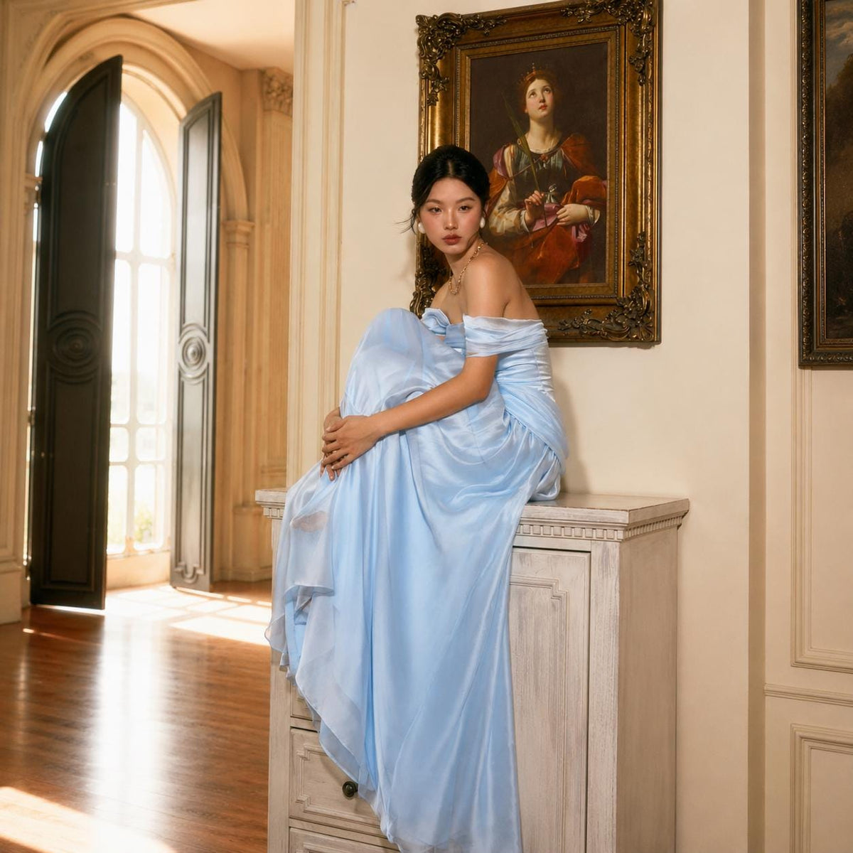 May include: A floor-length, light blue off-the-shoulder gown with a sweetheart neckline and a flowing, draped skirt. The dress features a smooth, satin-like finish and a fitted bodice that transitions into a voluminous, floor-sweeping hemline.