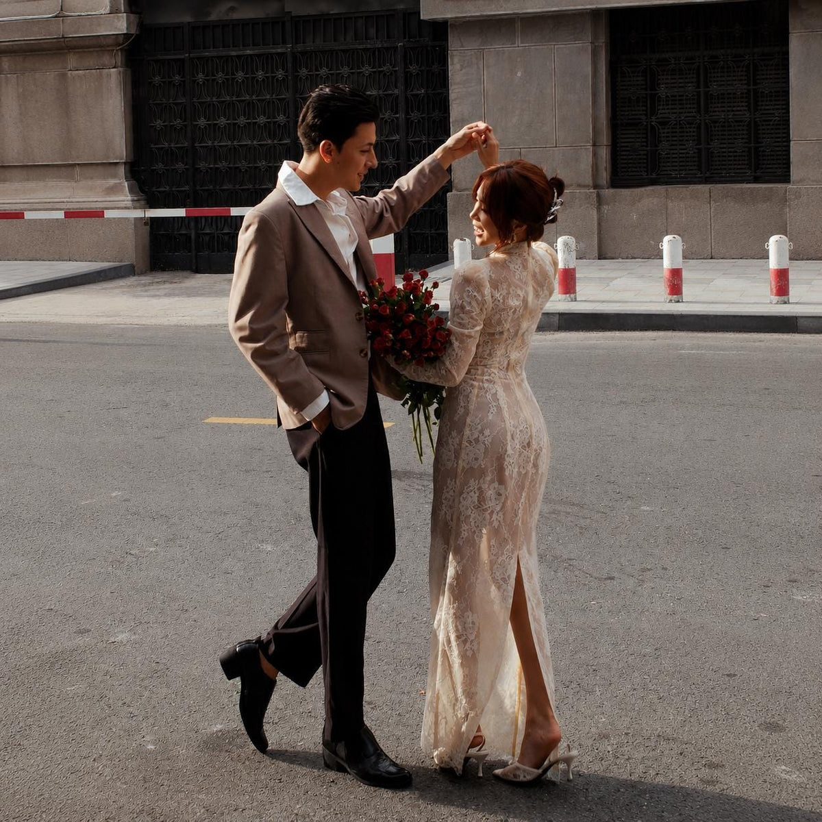 May include: A tan blazer and black trousers paired with a white collared shirt, alongside a floor-length cream lace dress with long sleeves and a back slit. A bouquet of deep red roses is held between the two outfits in an outdoor setting.