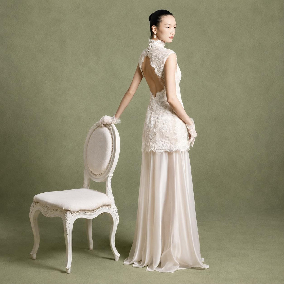 May include: A floor-length gown featuring a sleeveless, high-neck lace bodice with a teardrop-shaped open back and a row of small buttons at the nape. The skirt is made of a sheer, flowing fabric that drapes to the floor in a soft, neutral tone.