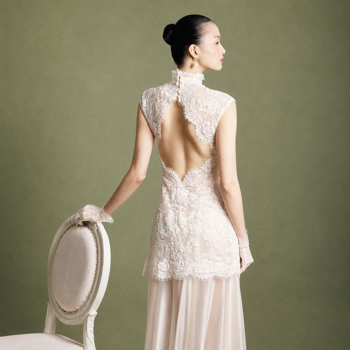 May include: A sleeveless ivory lace dress featuring a high collar with button closures, an open back design, and a sheer floor-length skirt. The bodice is adorned with intricate floral embroidery and lace detailing, paired with matching lace gloves.