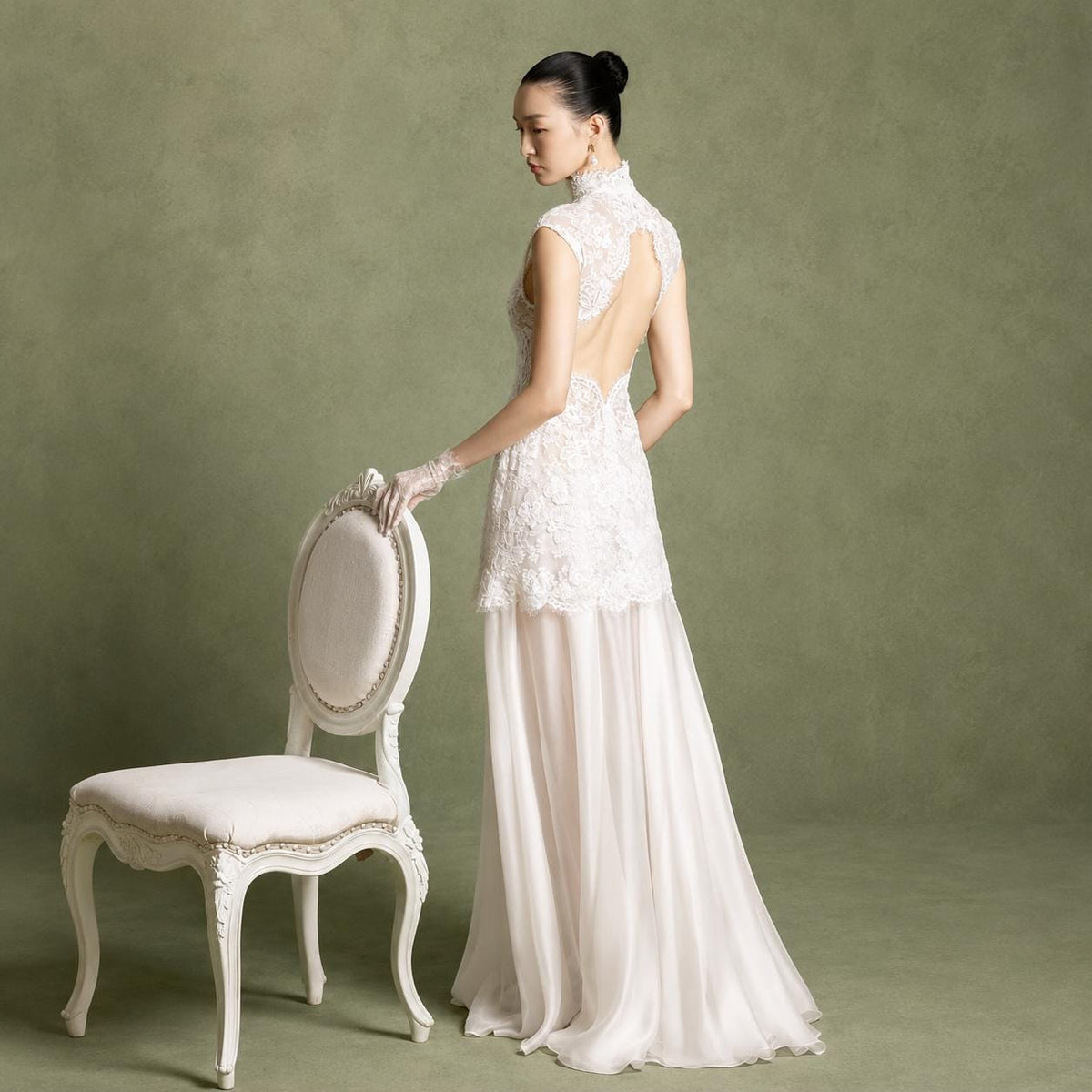 May include: A floor-length white gown featuring a high-neck lace bodice with an open back design, short cap sleeves, and a flowing sheer skirt. The garment is paired with a white wooden chair that has a rounded backrest and upholstered seat.