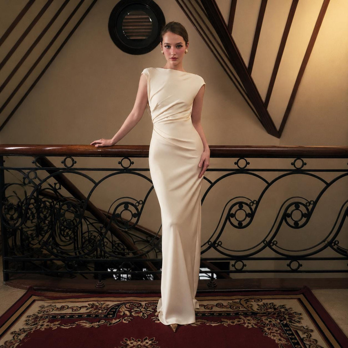 May include: A floor-length, cream-colored gown featuring a boat neckline, cap sleeves, and intricate side-waist ruching. The garment is crafted from a smooth, satin-like fabric that drapes down to the floor, creating a sleek and form-fitting silhouette.