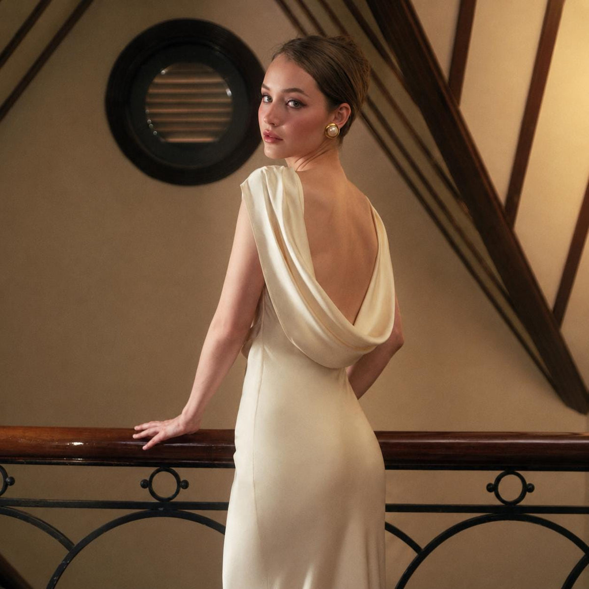 May include: A floor-length cream-colored satin dress featuring a draped cowl neckline at the back, sleeveless cut, and a smooth, flowing silhouette that falls straight to the floor. The fabric has a subtle sheen and a soft, light-reflecting finish.