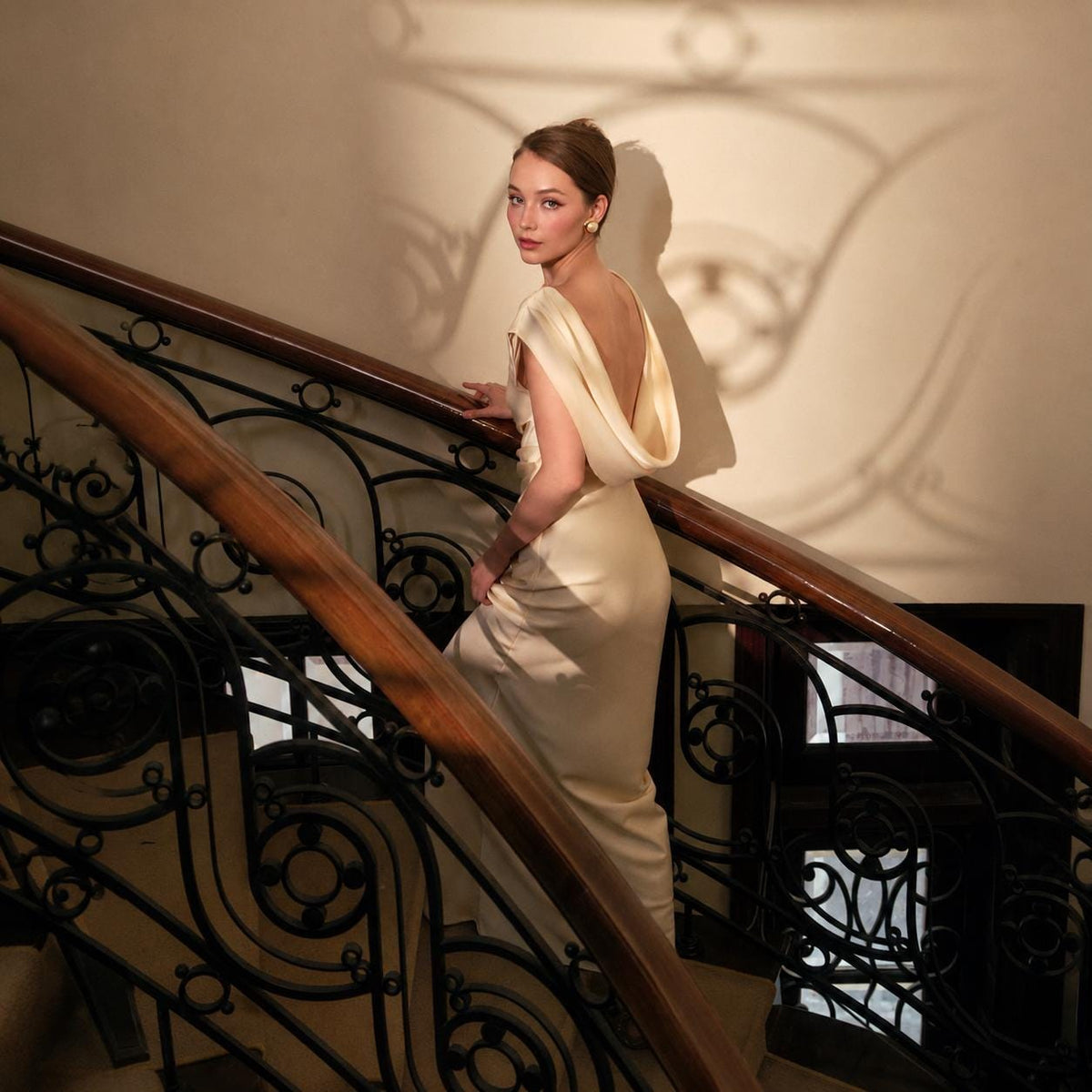 May include: A floor-length cream-colored gown featuring a cowl neckline and an open back design. The dress is sleeveless with a draped bodice and a long, straight skirt that falls to the floor, crafted from a smooth, light-reflecting fabric.