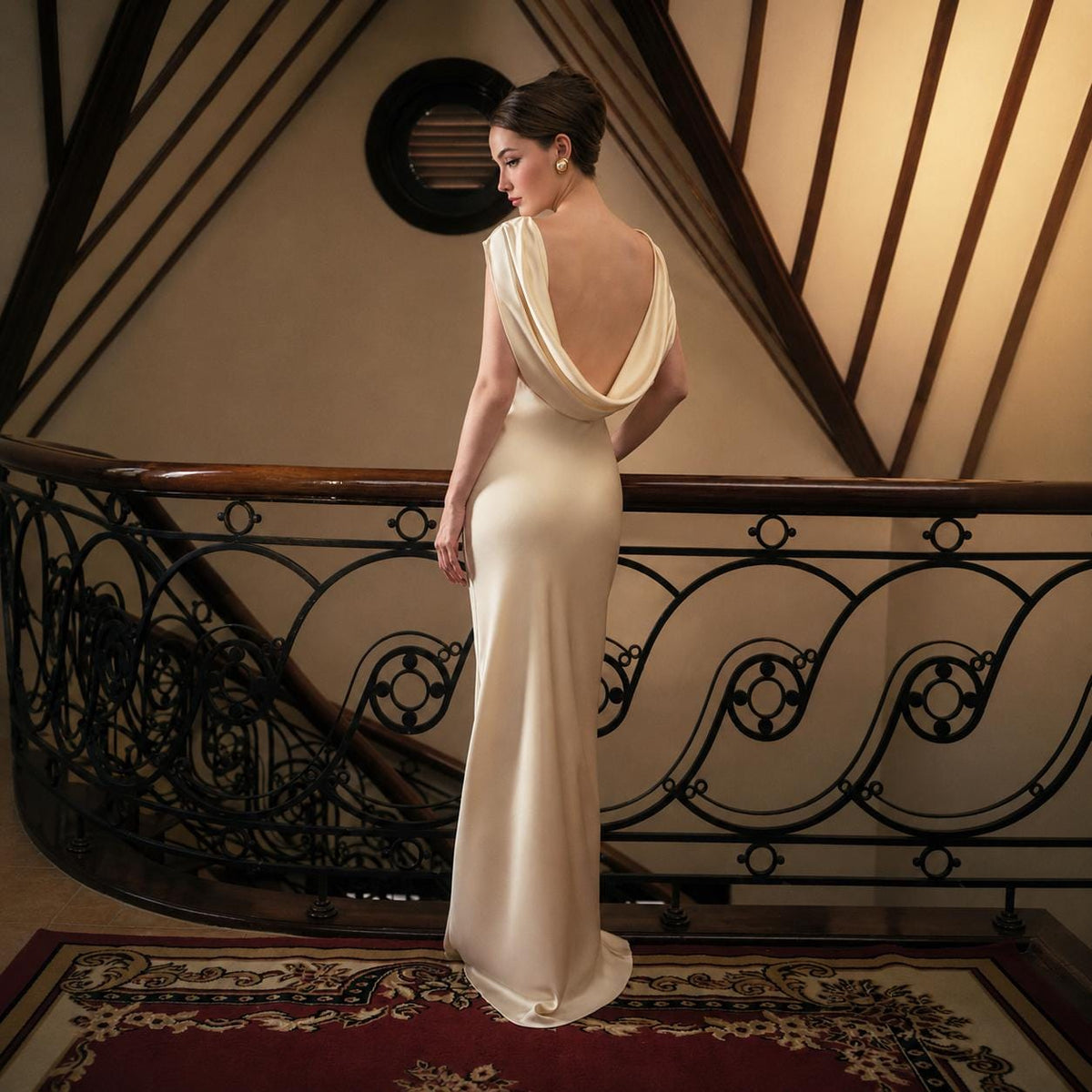 May include: A floor-length, cream-colored gown featuring a cowl neckline that drapes across the back, sleeveless shoulders, and a fitted silhouette that extends to the floor. The garment is made of a smooth, solid fabric with a subtle sheen.