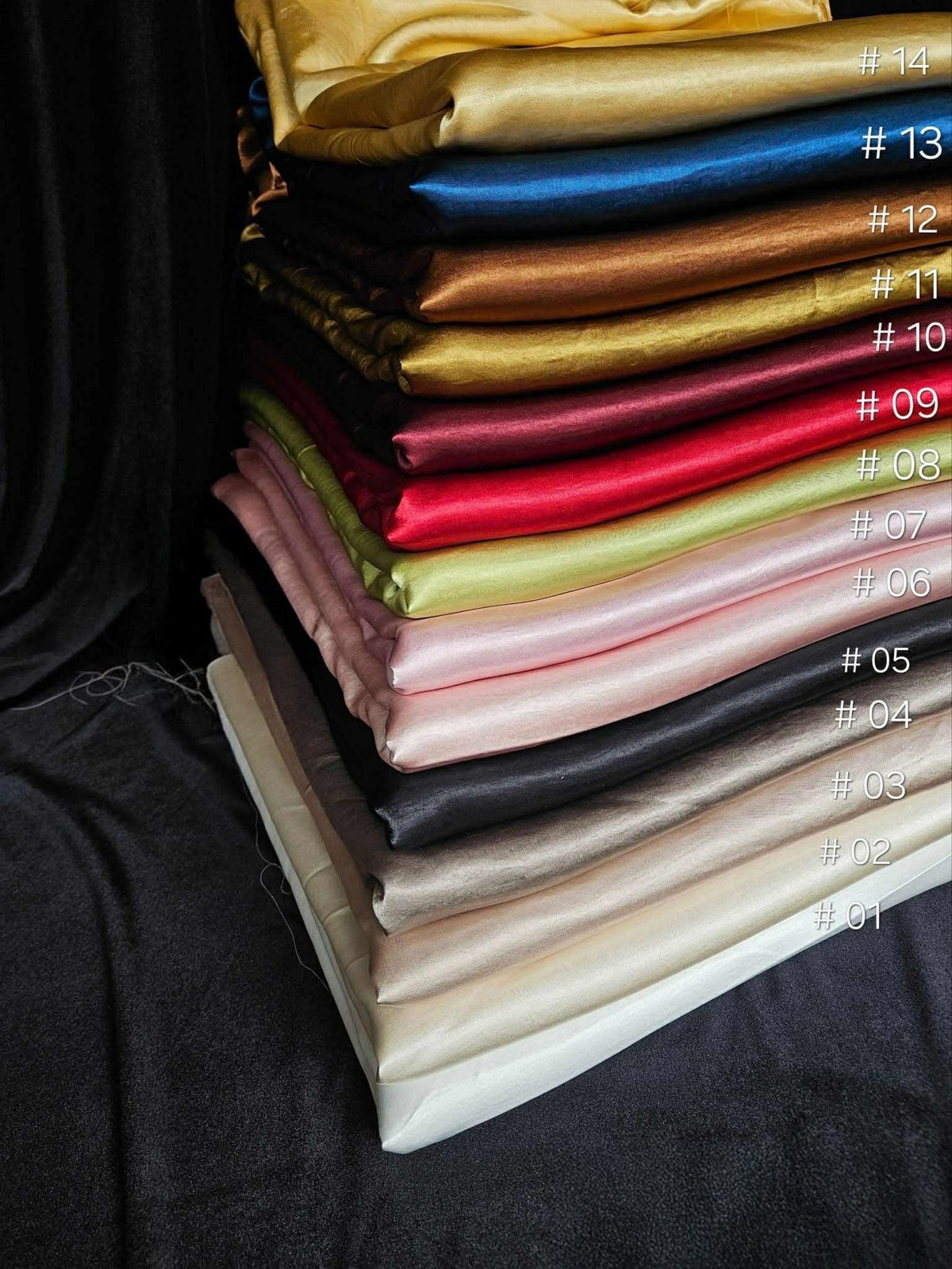 May include: A stack of folded satin fabric pieces in various colors, labeled from #01 to #14. The colors include white, beige, gray, black, light pink, pale green, red, dark red, gold, brown, blue, and yellow, all featuring a smooth, shiny finish.