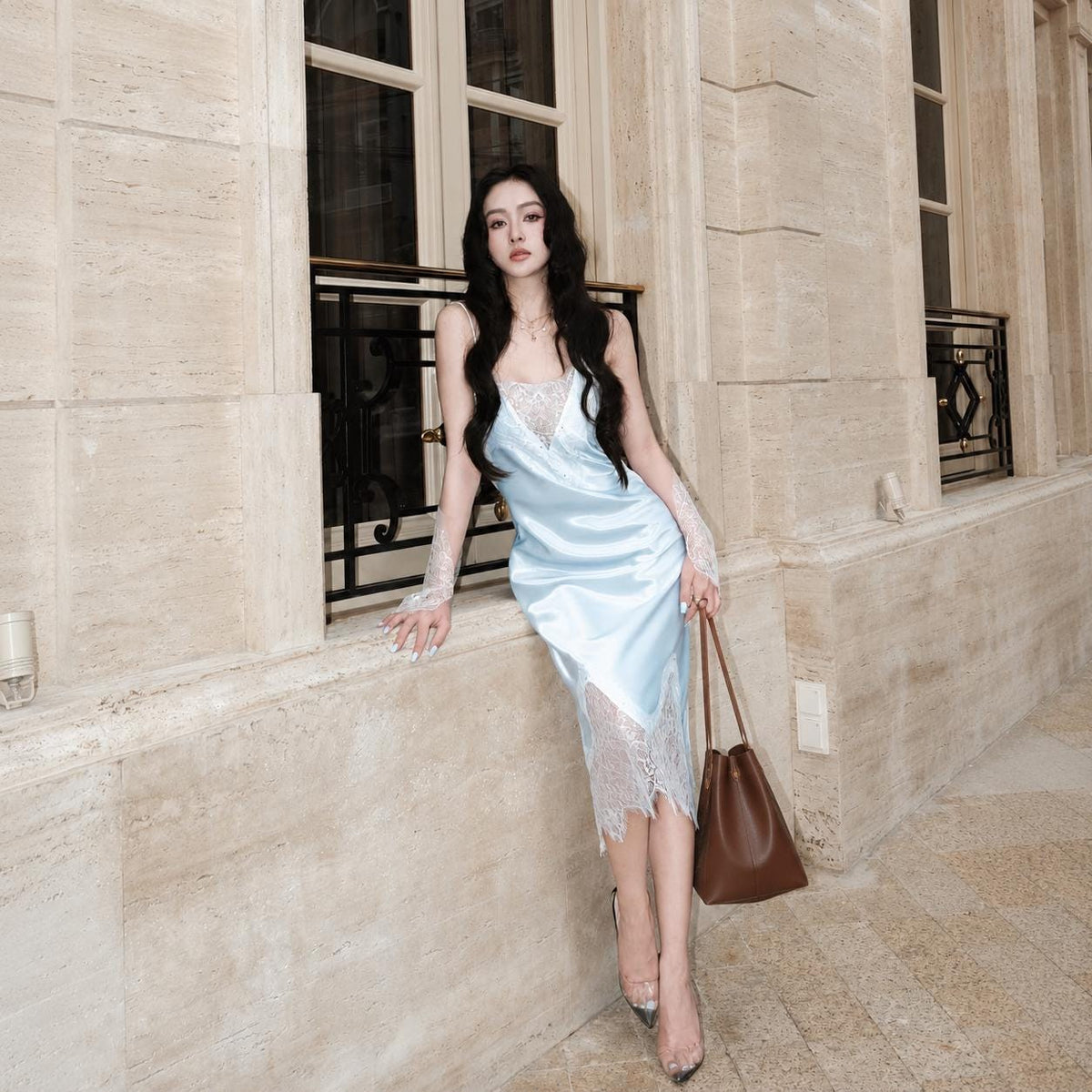 May include: A light blue satin midi dress featuring white lace trim along the neckline and a jagged lace hemline. The dress has thin spaghetti straps and is paired with a brown leather bucket bag and clear high-heeled shoes with pointed toes.