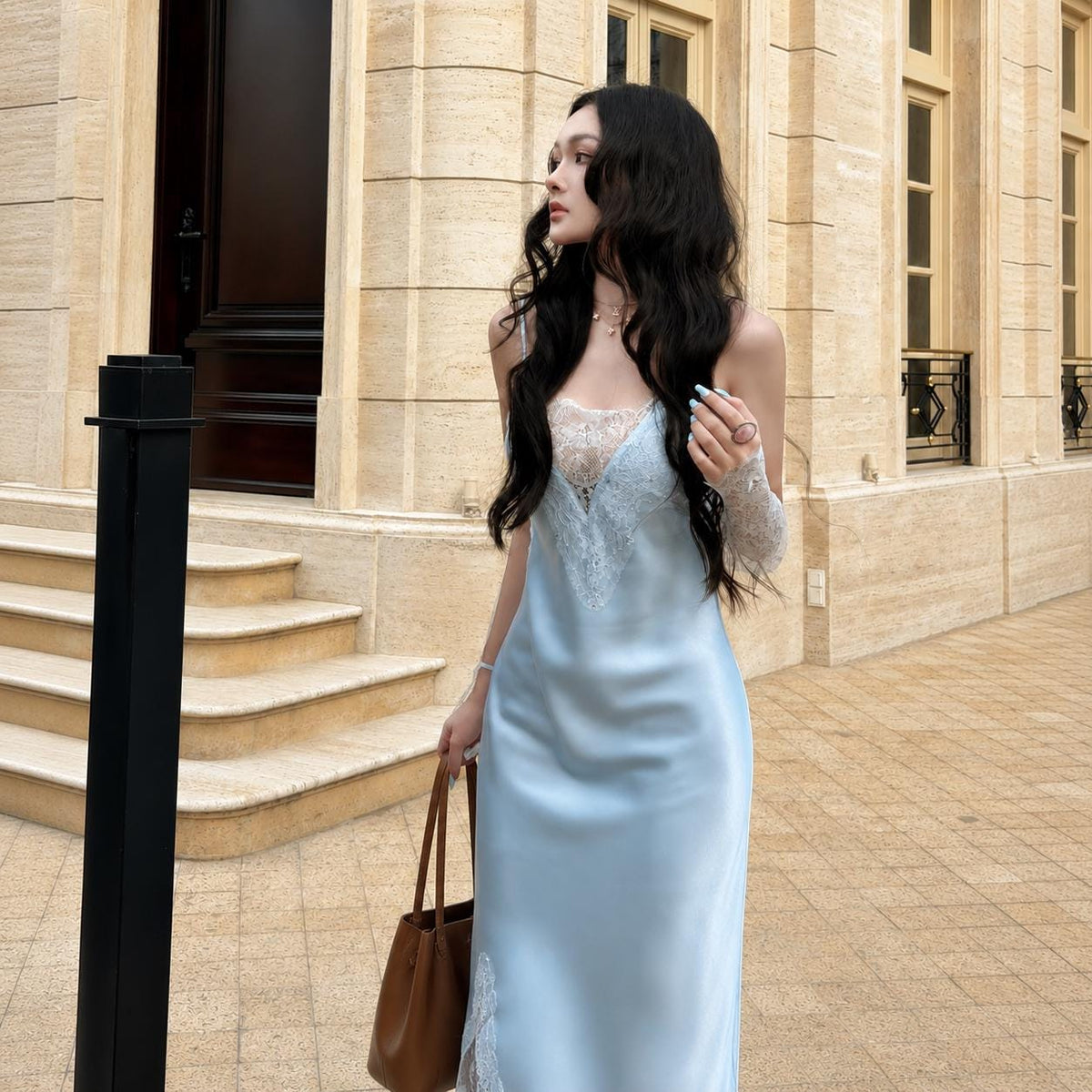 May include: A light blue satin slip dress featuring a V-neckline with white lace trim, thin spaghetti straps, and a matching lace detail at the side hem. The garment is paired with sheer lace arm sleeves and a brown leather tote bag.