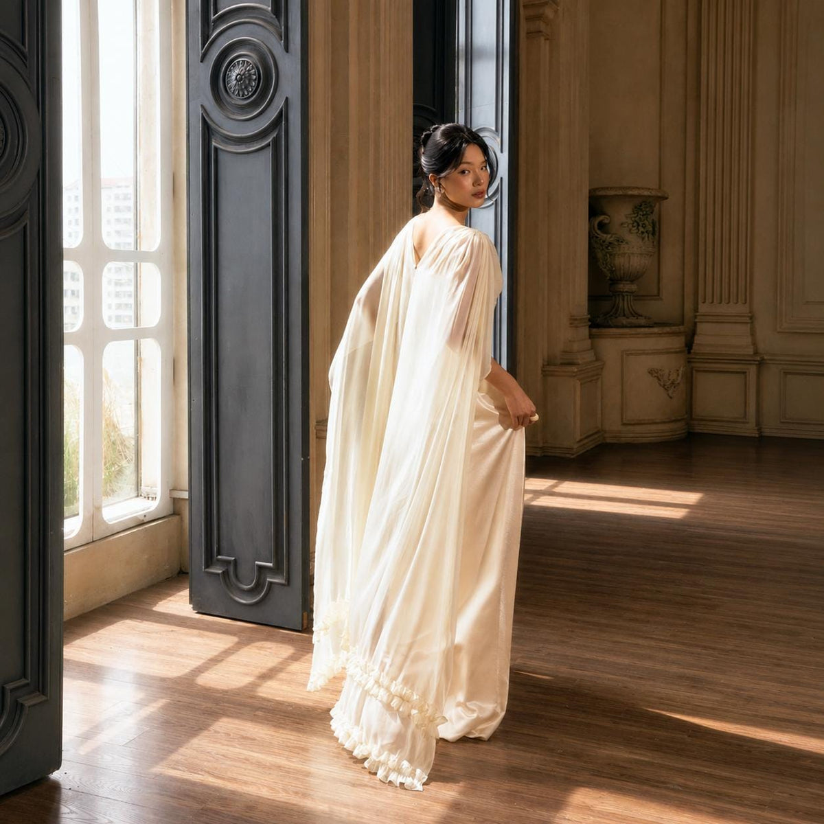 May include: A floor-length cream-colored gown featuring a flowing cape-style back, a sleeveless bodice, and a skirt with a ruffled hemline. The garment is crafted from a smooth, lightweight fabric that drapes down to the floor in soft folds.