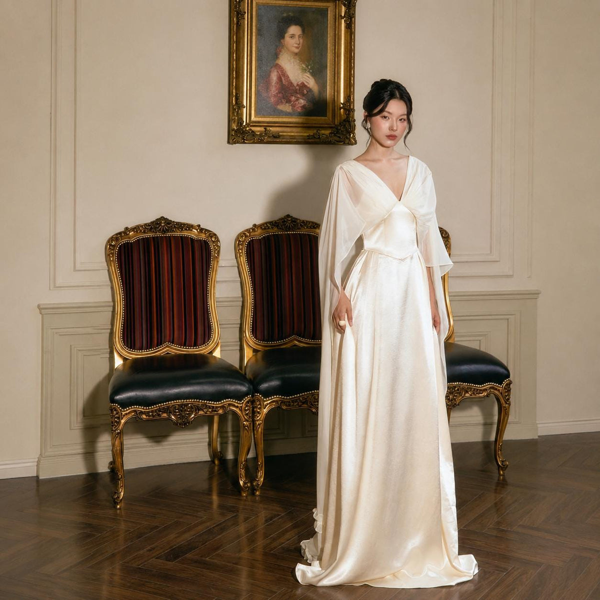 May include: A floor-length cream-colored satin gown featuring a V-neckline and flowing cape-style sleeves. The dress has a fitted bodice that transitions into a smooth, floor-length skirt, crafted from a lustrous fabric with a subtle sheen.