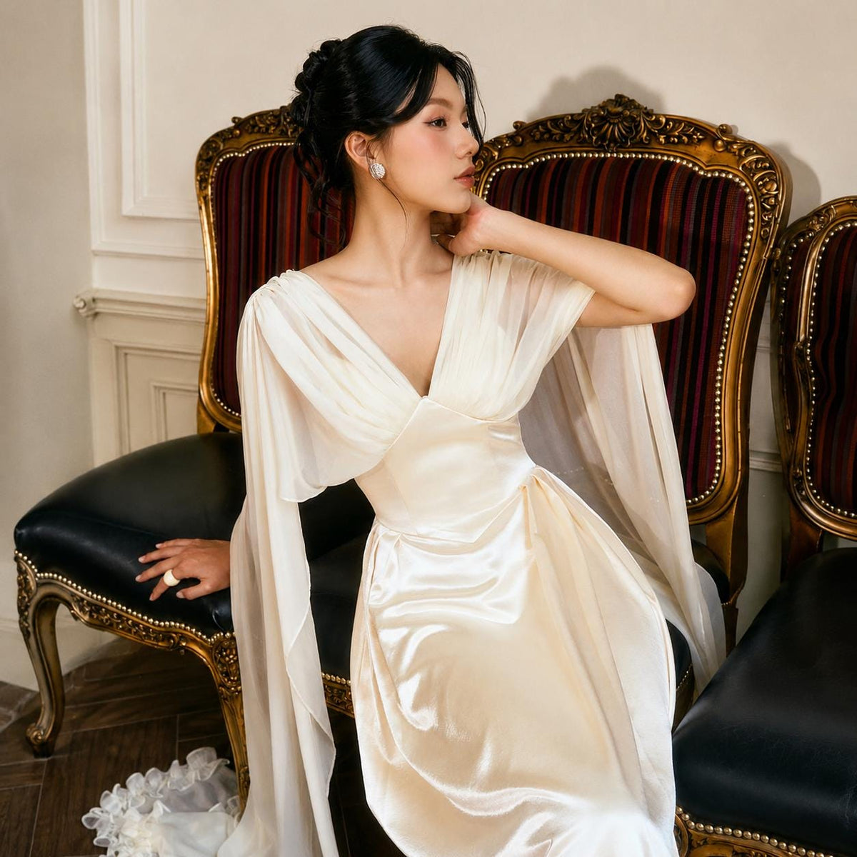 May include: A floor-length cream-colored satin gown featuring a deep V-neckline, a fitted bodice, and long, flowing sheer chiffon sleeves that drape from the shoulders. The dress has a smooth, lustrous finish and a soft, elegant silhouette.