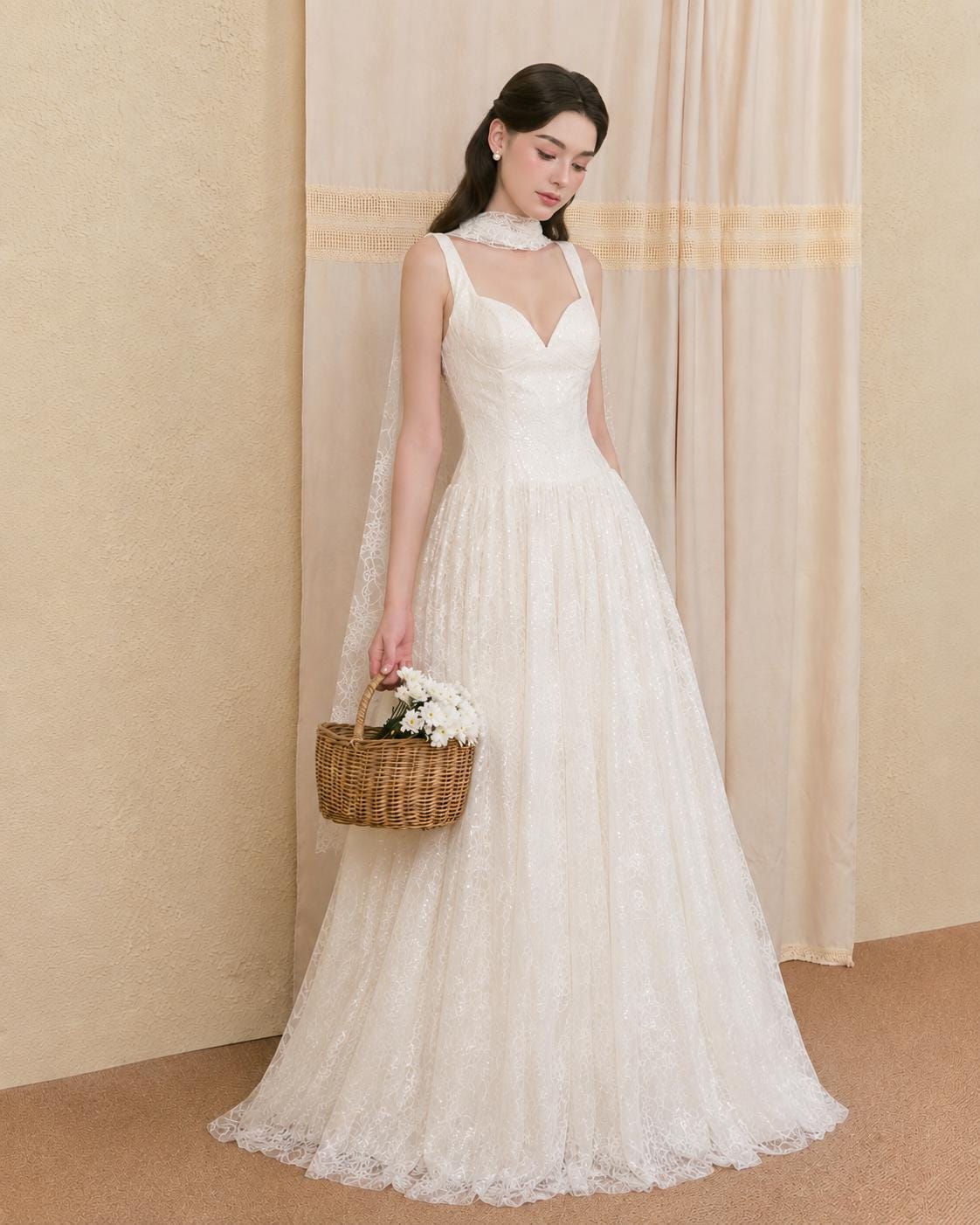 May include: A floor-length white lace gown featuring a sweetheart neckline, thick shoulder straps, and a fitted bodice that transitions into a full, flowing skirt. A matching lace neck scarf and a small brown woven basket with white flowers.