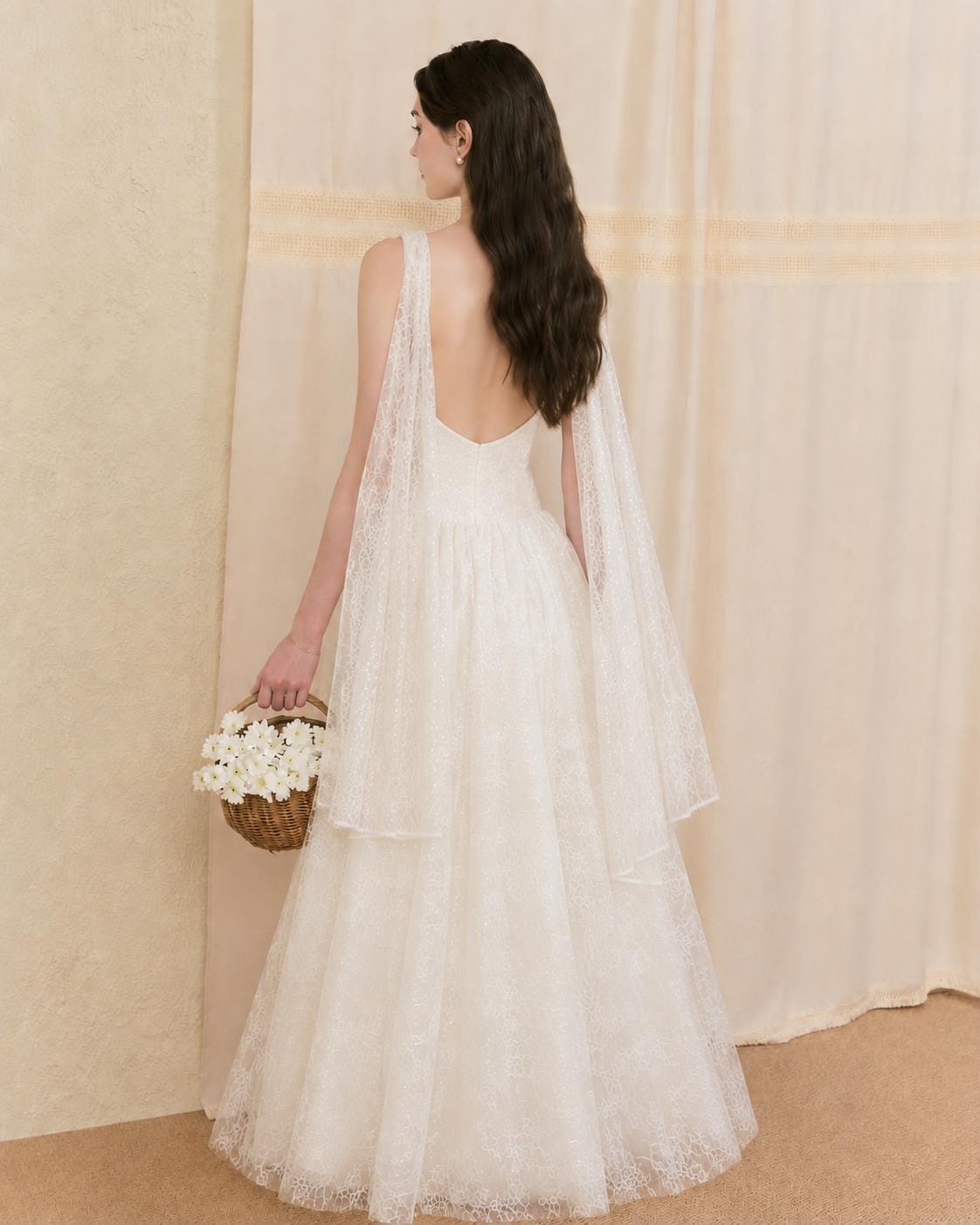 May include: A floor-length white lace dress featuring a deep scoop back, sleeveless bodice, and flowing sheer lace cape-style sleeves that drape from the shoulders. The dress has a full skirt and is paired with a small brown woven basket of white flowers.