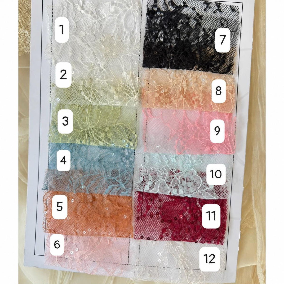 May include: A color swatch card featuring twelve distinct samples of floral lace fabric, each numbered from one to twelve. The lace swatches display a variety of colors including white, black, green, blue, orange, pink, and red, with visible sequins.