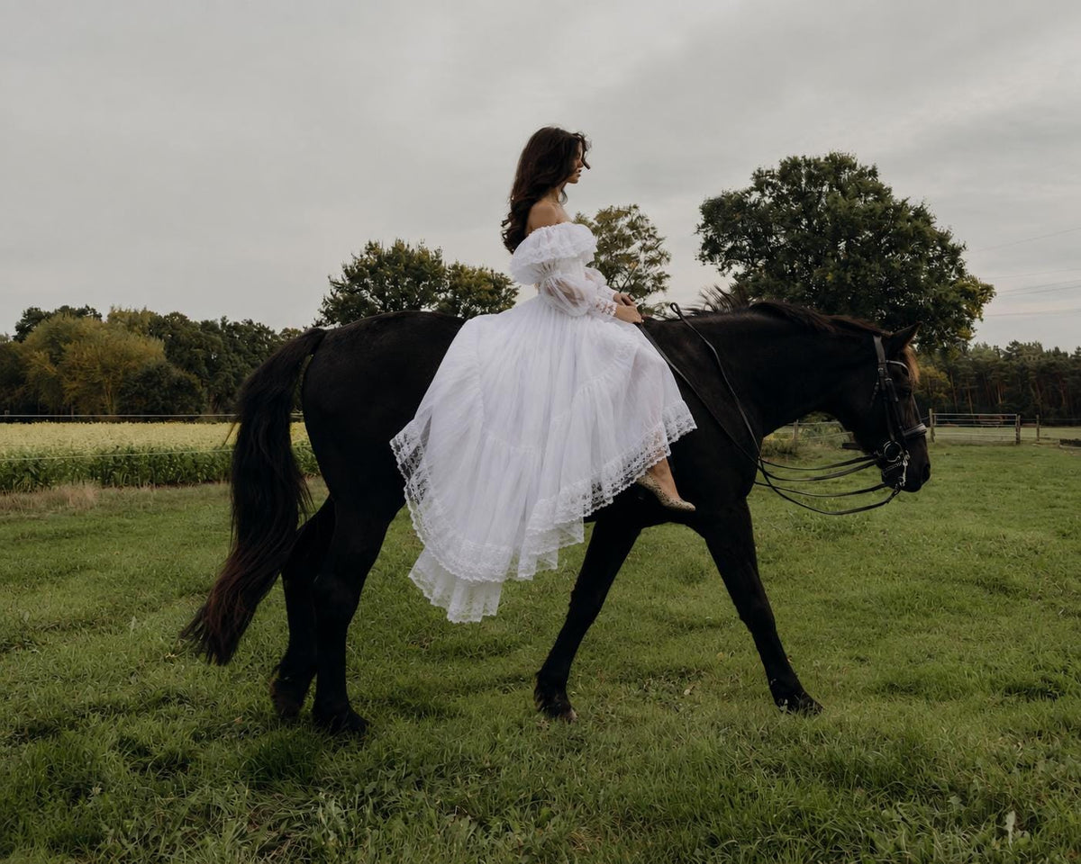 May include: A white off-the-shoulder dress with long sheer sleeves and a layered skirt featuring lace trim details. The garment is worn by a person riding a black horse in a grassy field under an overcast sky, showcasing the full-length design.