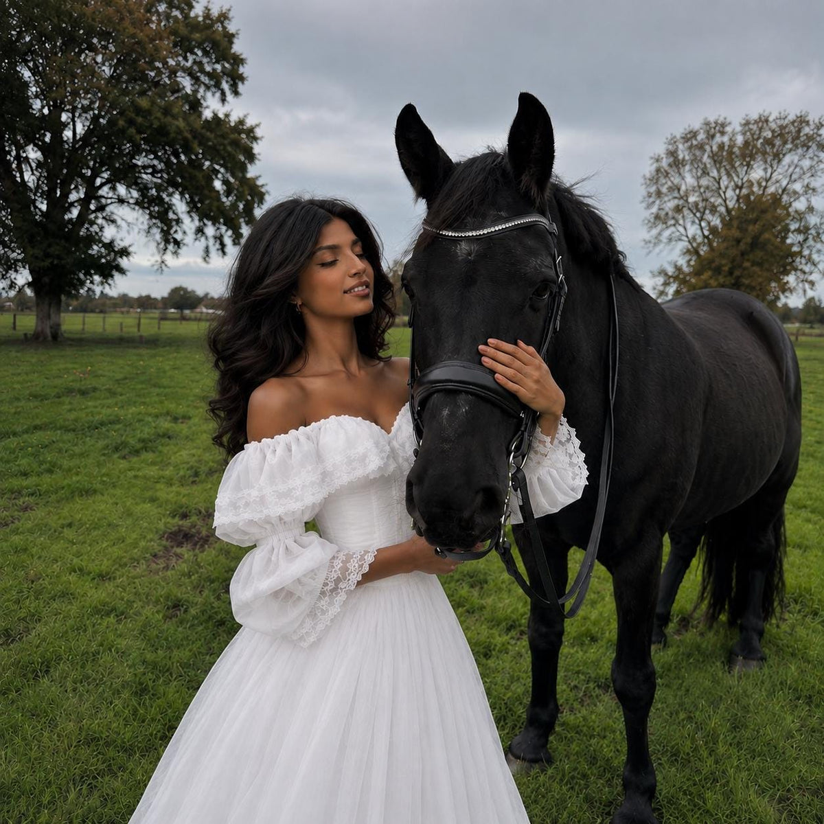 May include: A white off-the-shoulder gown featuring a ruffled neckline, long sheer sleeves with lace trim, and a full-length skirt. The dress is made of a lightweight, layered fabric and is paired with a black horse wearing a leather bridle.