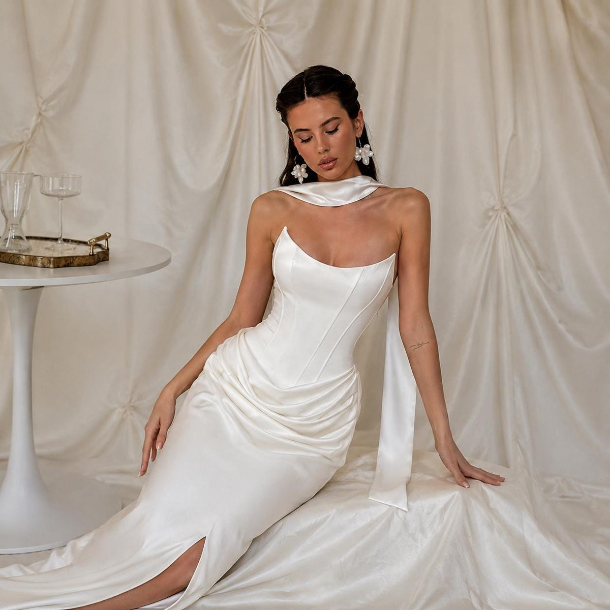 May include: A floor-length ivory satin gown featuring a structured corset-style bodice with a scoop neckline, a draped skirt with a side slit, and a matching neck scarf. The garment is crafted from a smooth, lustrous fabric with visible seams.