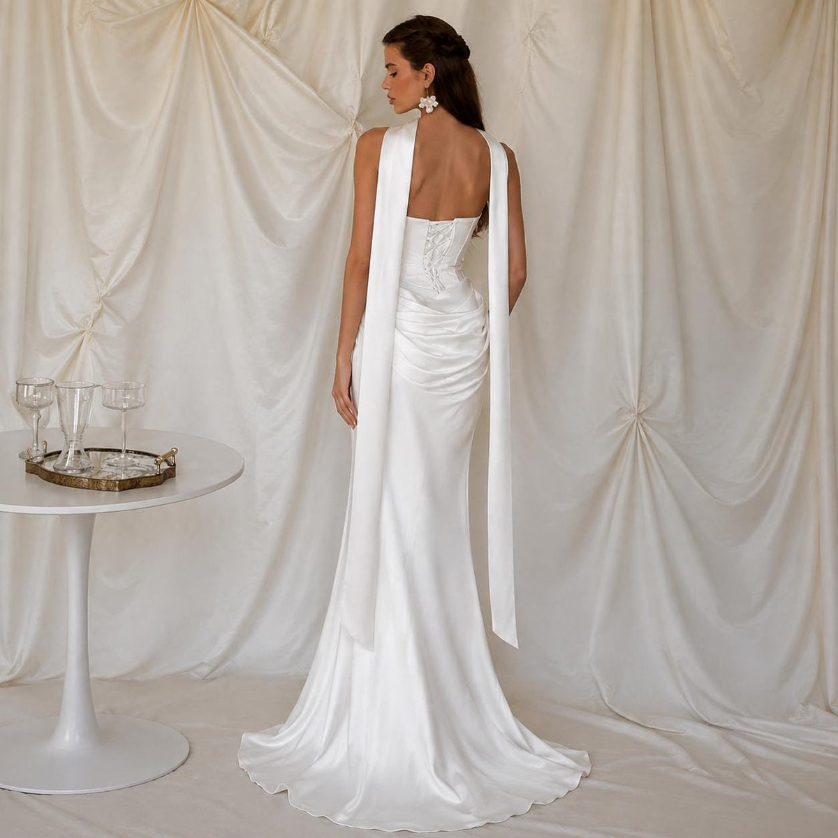 May include: A floor-length white satin gown featuring a strapless bodice with a corset-style back, a draped waistline, and two long, flowing fabric panels extending from the shoulders down the back, creating a sleek and elegant silhouette.