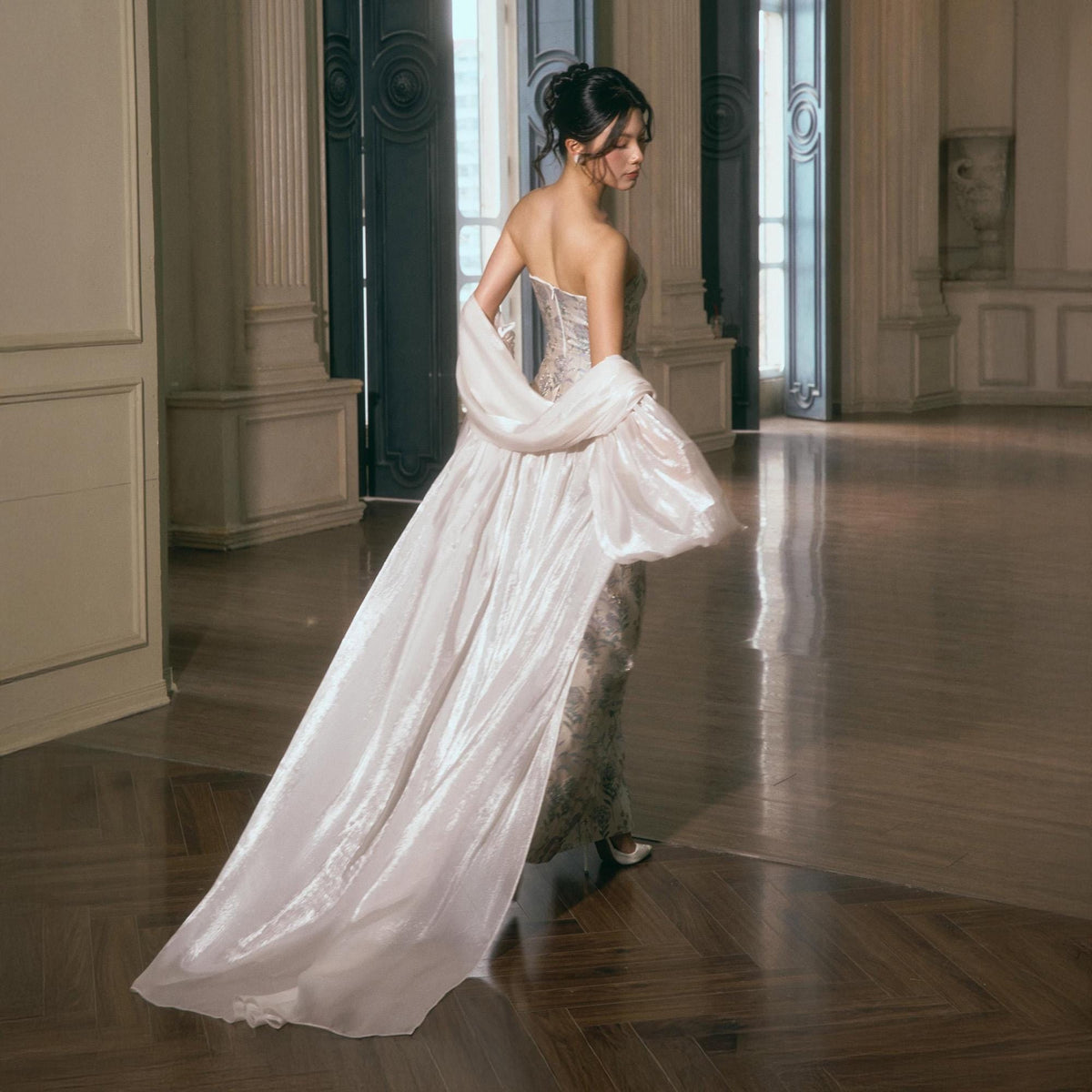 May include: A strapless floor-length gown featuring a subtle floral pattern in muted tones, paired with a long, sheer, iridescent white shawl that drapes over the shoulders and extends to the floor, creating a flowing silhouette with a train.