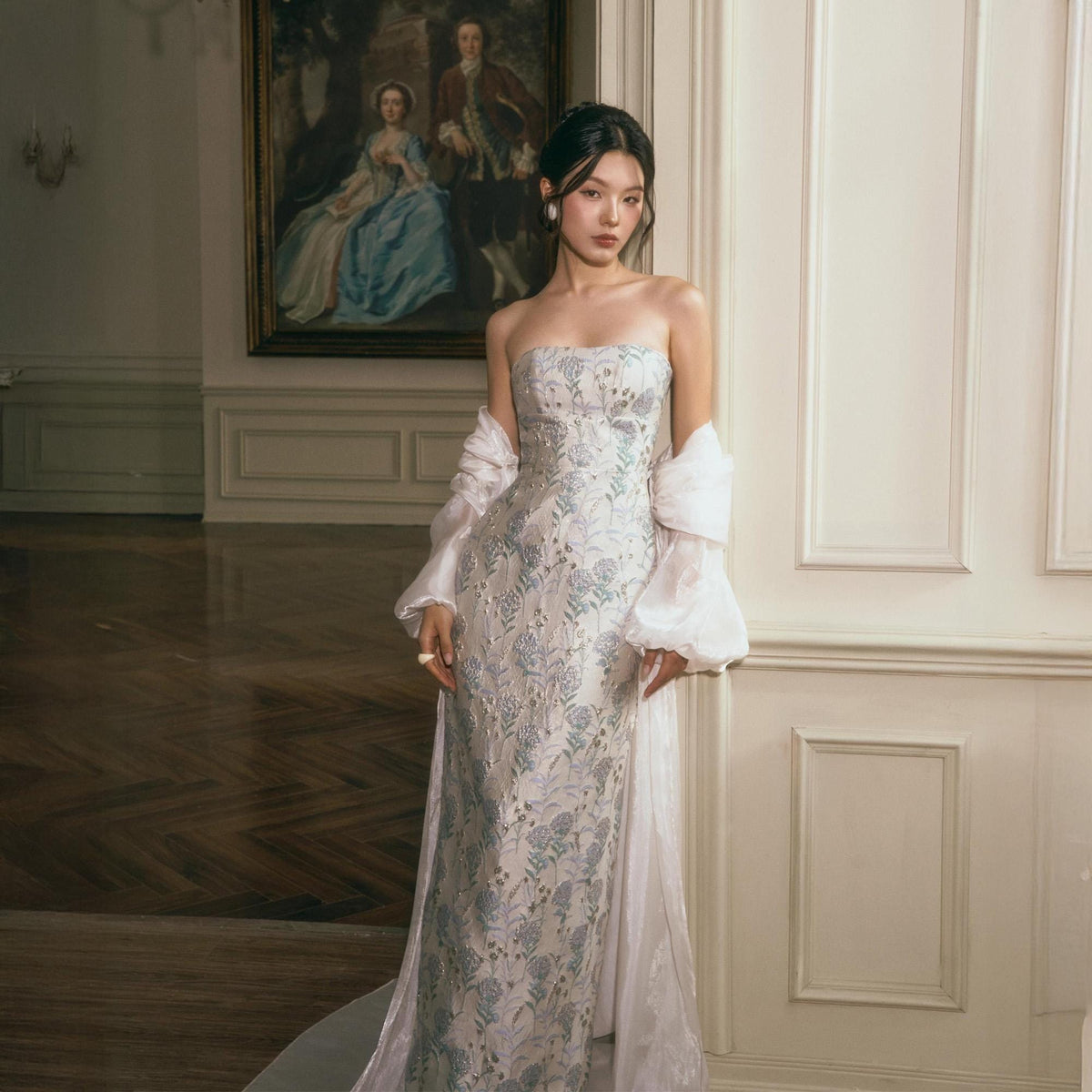 May include: A strapless floor-length gown featuring a light-colored fabric with an intricate blue and silver floral pattern. The dress is paired with sheer, voluminous, long-sleeved white bolero-style sleeves that drape loosely over the arms.