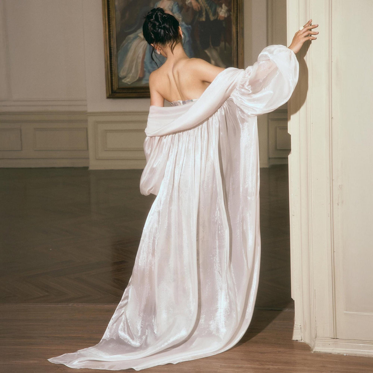 May include: A floor-length, off-the-shoulder robe in a shimmering, pale iridescent fabric. The garment features voluminous, sheer bishop sleeves and a flowing, loose-fitting silhouette that drapes to the floor, creating a soft, ethereal appearance.
