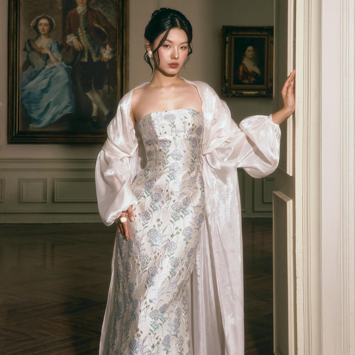 May include: A strapless floor-length gown featuring a white base with a light blue and silver floral pattern, paired with a matching sheer, long-sleeved, open-front duster coat. The ensemble is worn with a single white pearl earring and a ring.