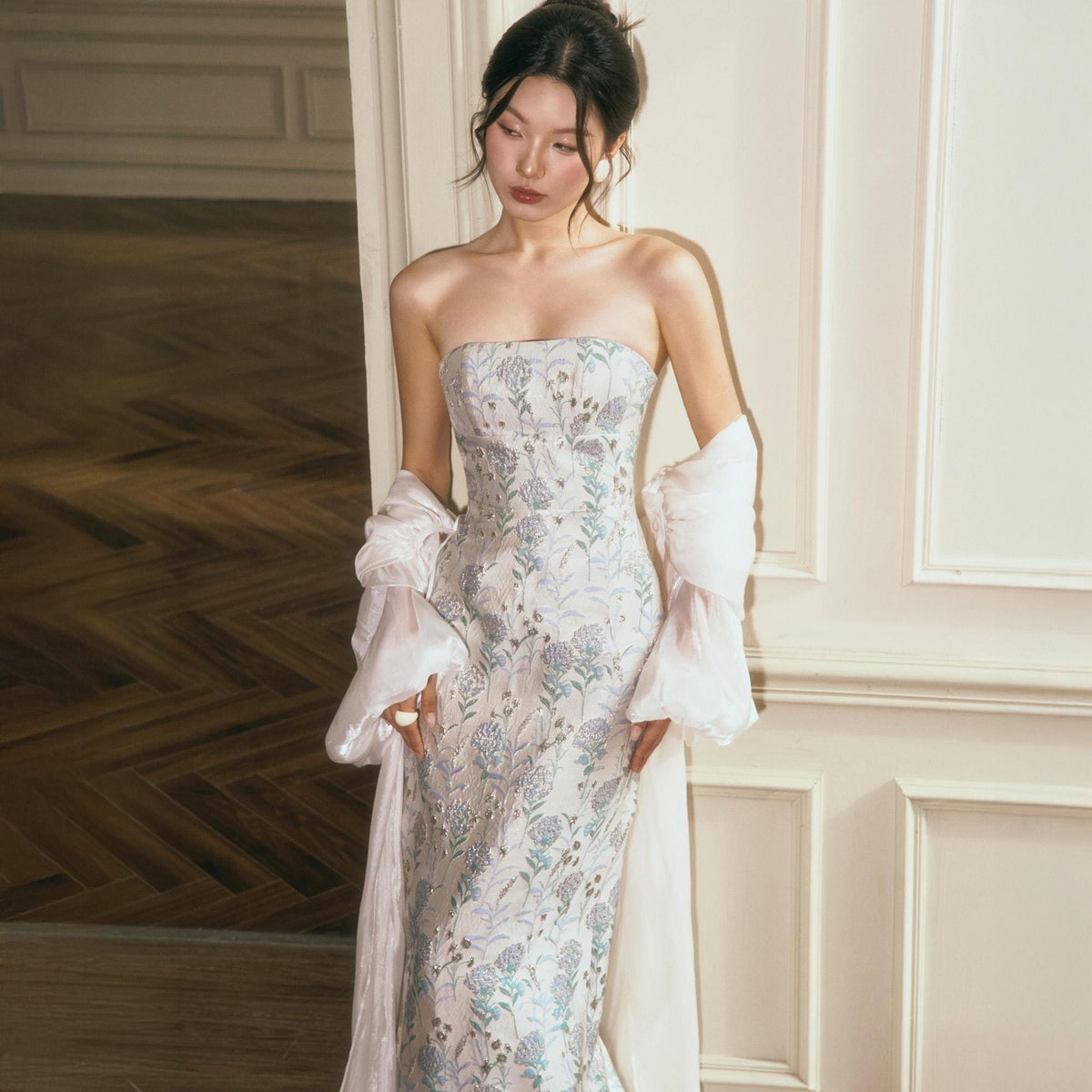 May include: A strapless floor-length gown featuring a light grey base with an intricate floral pattern in shades of blue, green, and silver. The dress is paired with sheer, voluminous off-the-shoulder sleeves that drape down the sides of the garment.