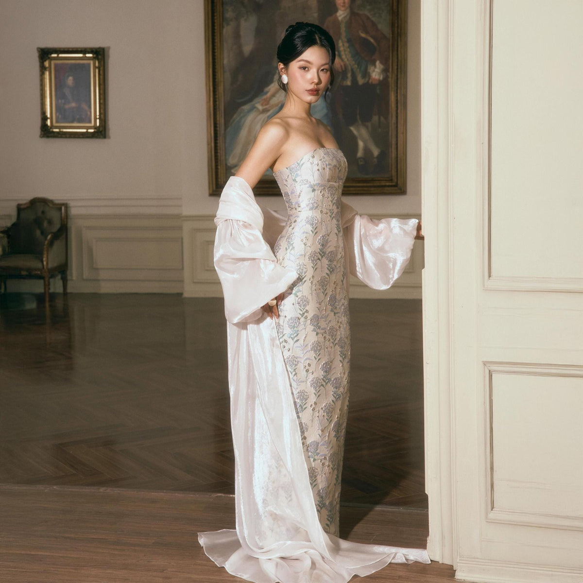 May include: A strapless floor-length gown featuring a light beige base with a blue floral pattern, paired with a matching sheer, long-sleeved, flowing wrap that drapes from the shoulders to the floor, creating a layered and elegant silhouette.