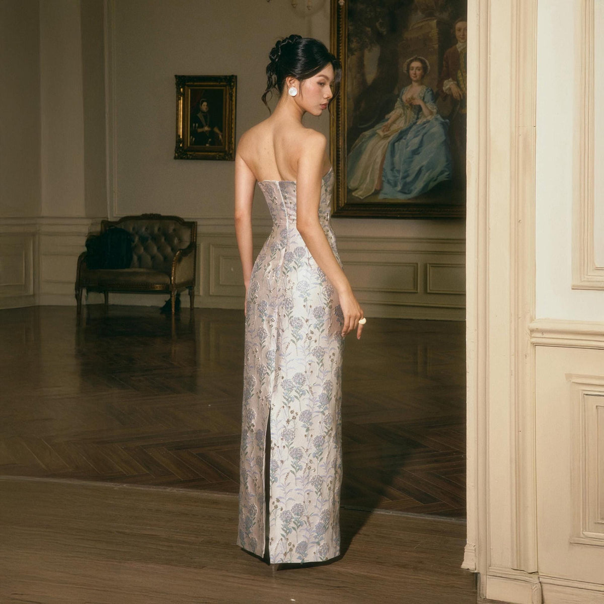 May include: A floor-length strapless dress featuring a light-colored fabric with a repeating floral pattern in shades of blue and green. The garment has a fitted bodice, a straight silhouette, and a vertical slit at the center back hemline.